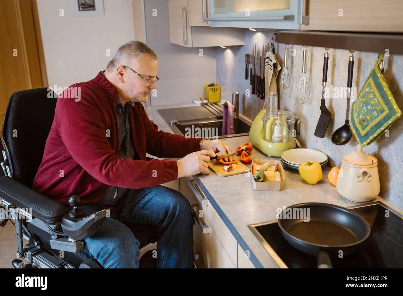 Retired senior man with disability sitting in motorized wheelchair cutting bell pepper at kitchen counter Stock Photo