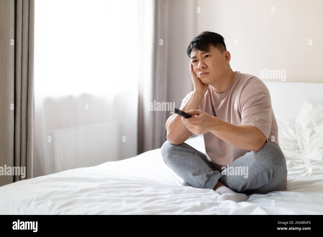 bored asian man sitting on bed at home, channels on tv Stock Photo
