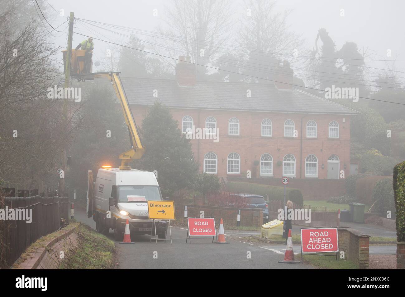 Telecommunications people working on overhead telephone lines from a cherry picker cradle vehicle. Resident asks questions of the worker in the fog. Stock Photo