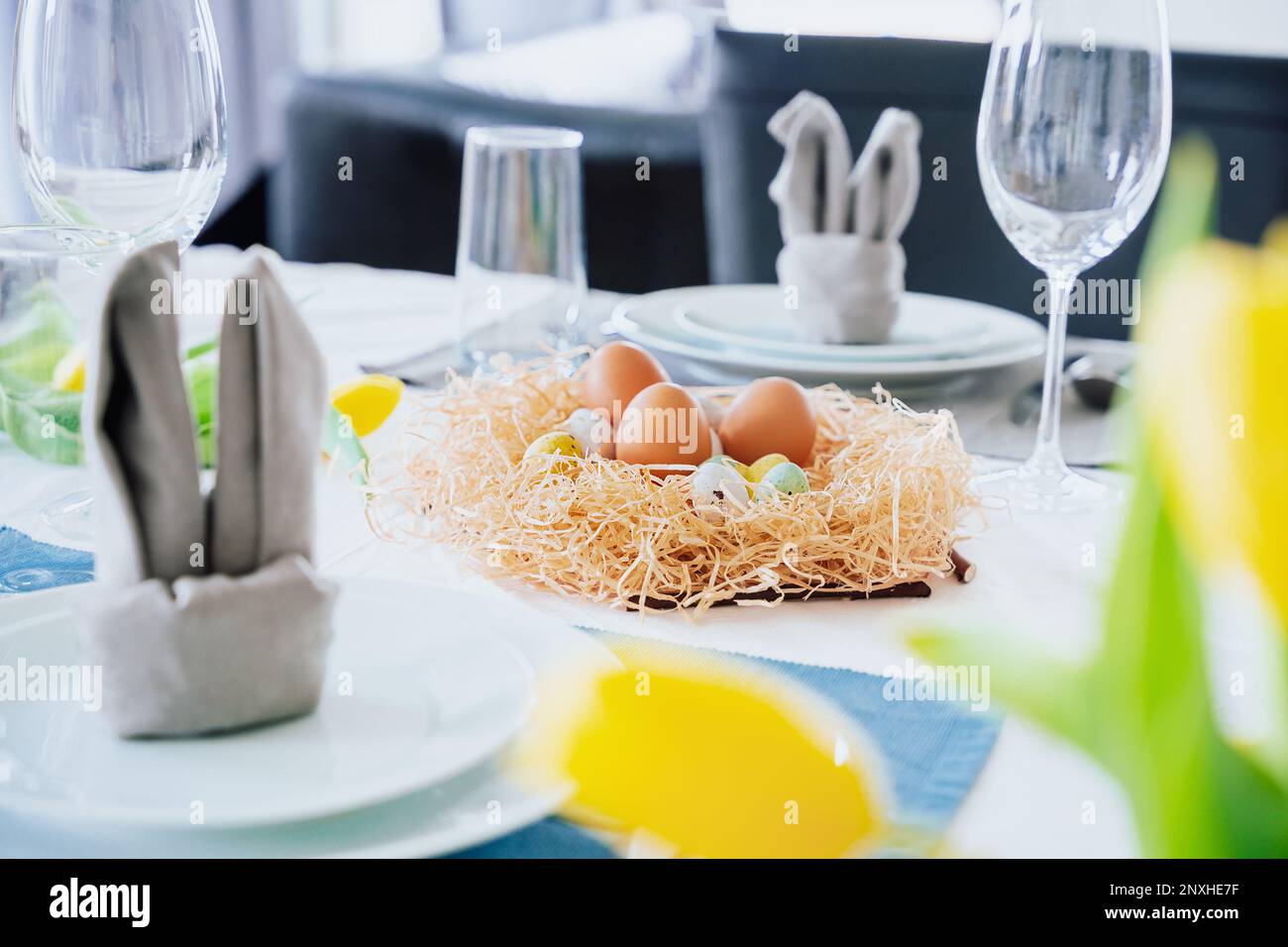 Beautiful Easter table setting with festive decor indoors. Napkin in shape of rabbit ears, colorful eggs in nest, fresh yellow tulips in vase. Sunligh Stock Photo