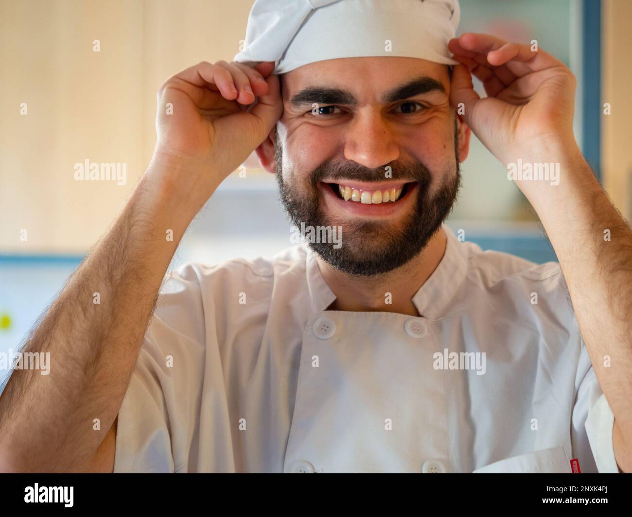 portrait of a young bearded chef smiling  while putting on his chef hat Looking at camera Stock Photo