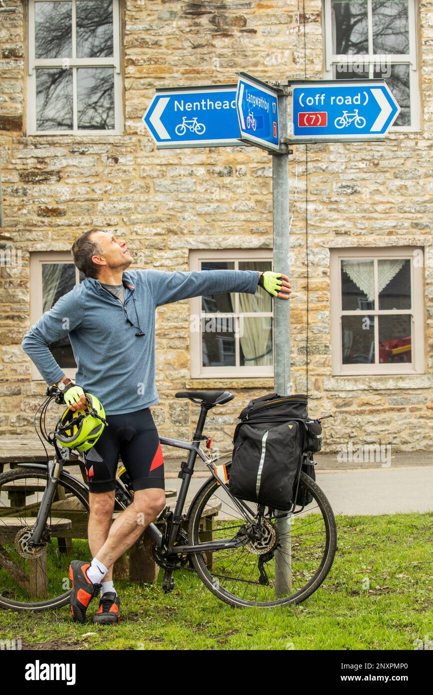 A cyclist with yellow helmet beneath a signpost for cycle routes to Nenthead, Langwathby and off road at Garrigill, Cumbria Stock Photo