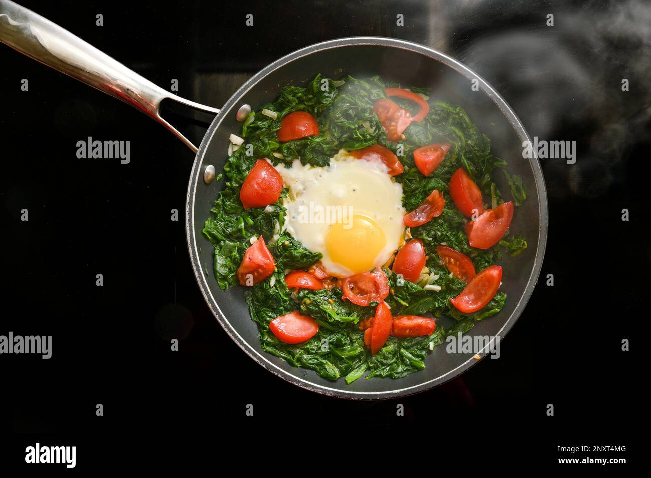 Spinach, tomatoes and fried egg in a steaming pan on a black stovetop, cooking a healthy meal for breakfast or lunch, high angle view from above, copy Stock Photo