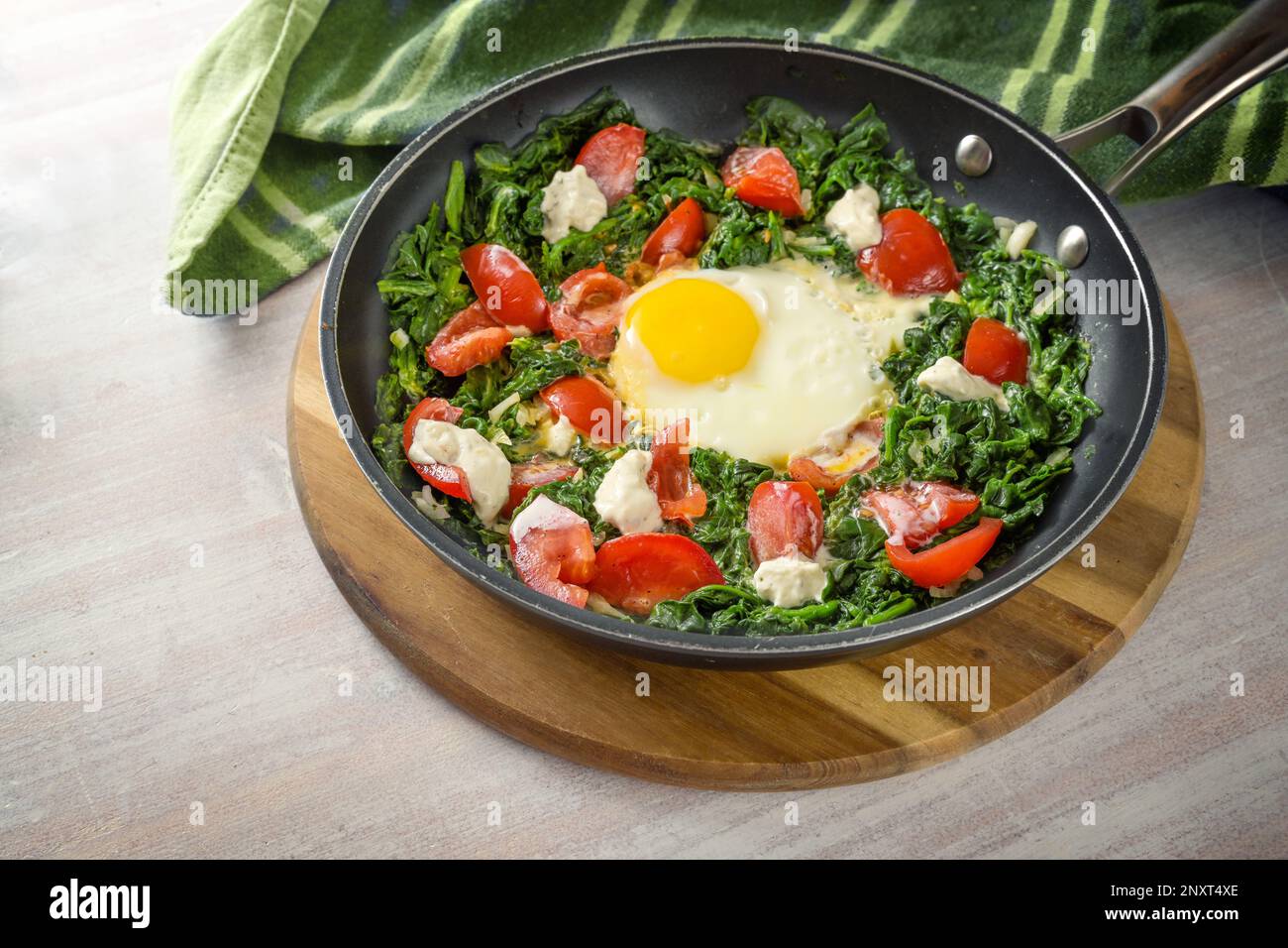 Fried egg, spinach and tomatoes with some yoghurt blobs in a frying pan on a wooden cutting board and a green kitchen towel, healthy meal, copy space, Stock Photo