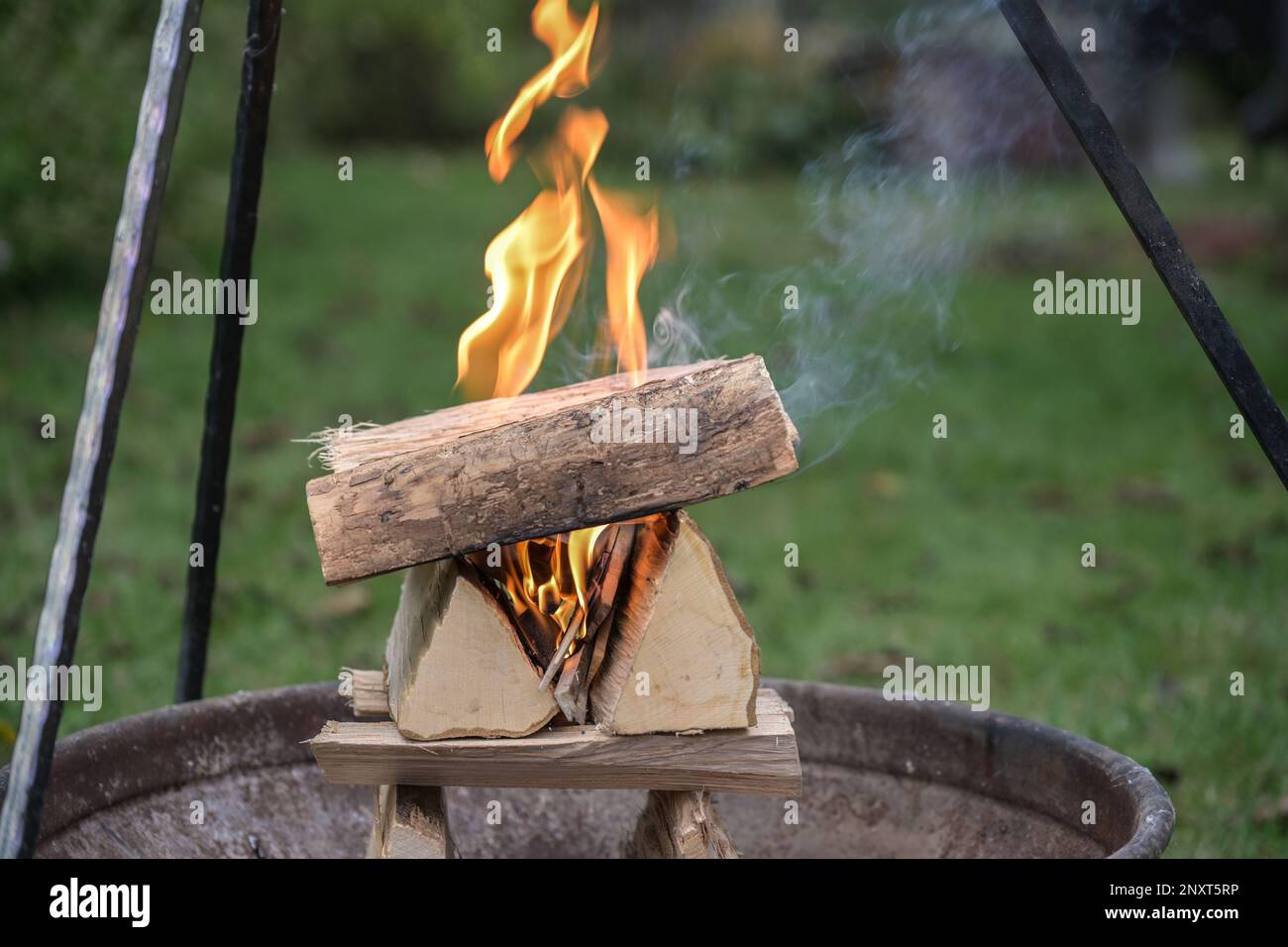 Clever stacked firewood logs burning with flames and smoke in a fire bowl under a tripod grill in a garden for a barbecue party, copy space, selected Stock Photo