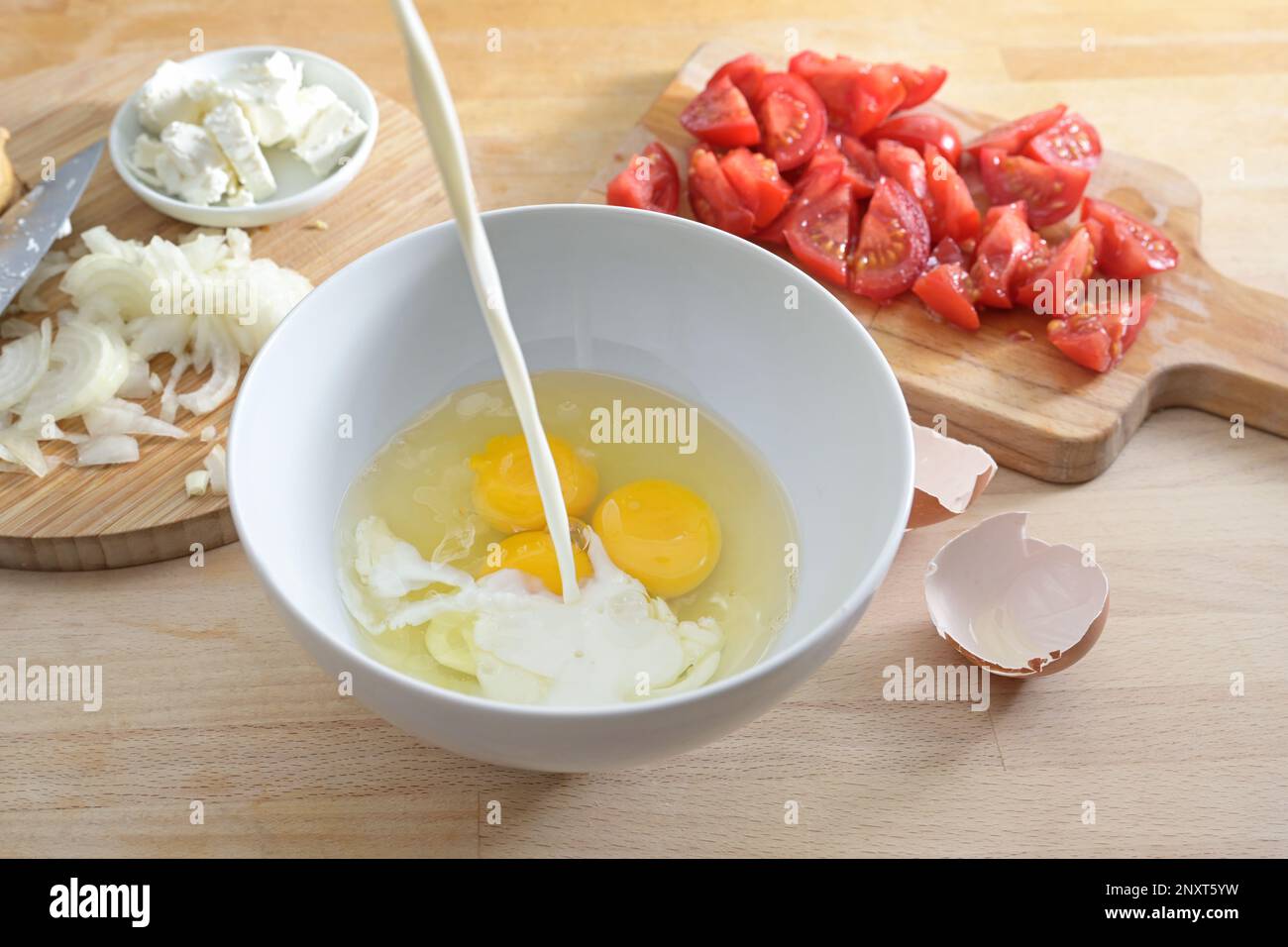 Pouring milk into a bowl with eggs, preparation for a frittata or omelet with tomatoes and onions, wooden kitchen board, selected focus, narrow depth Stock Photo