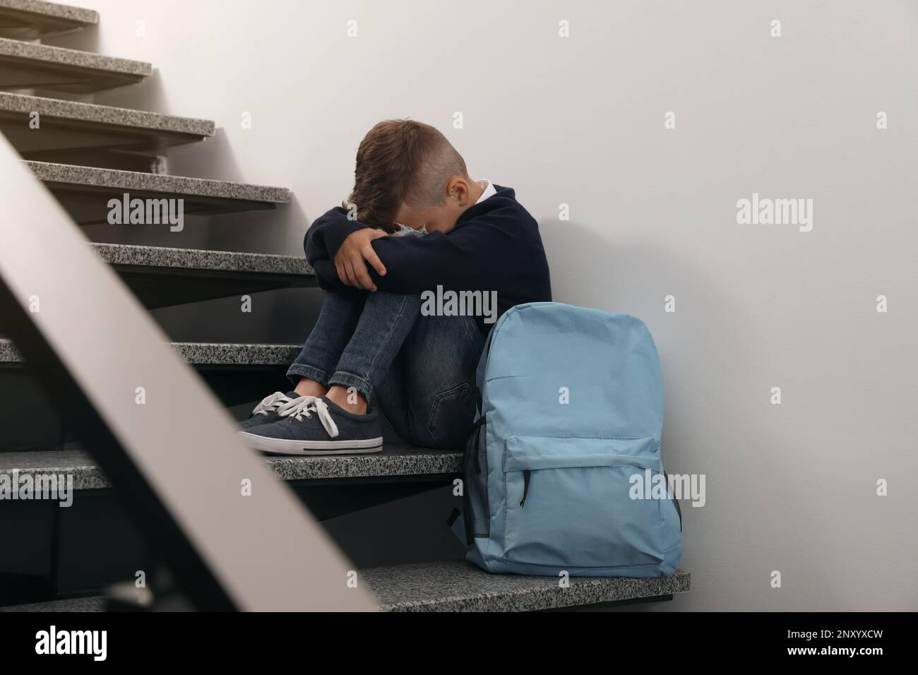 Upset little boy with backpack sitting on stairs indoors Stock Photo