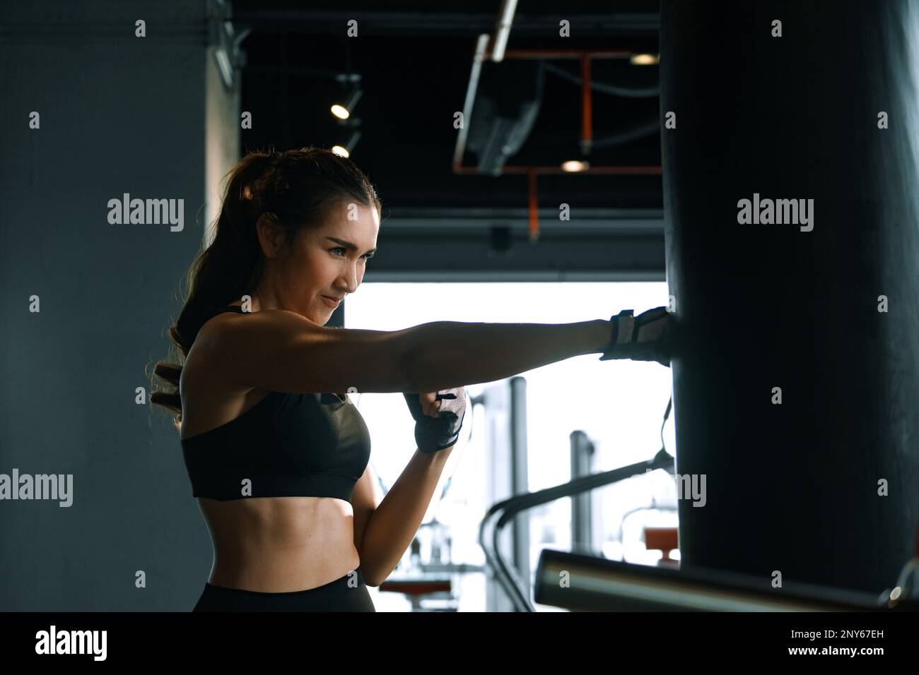 Young woman practicing boxing at the gym, she wears boxing gloves and hits a punching bag. Stock Photo