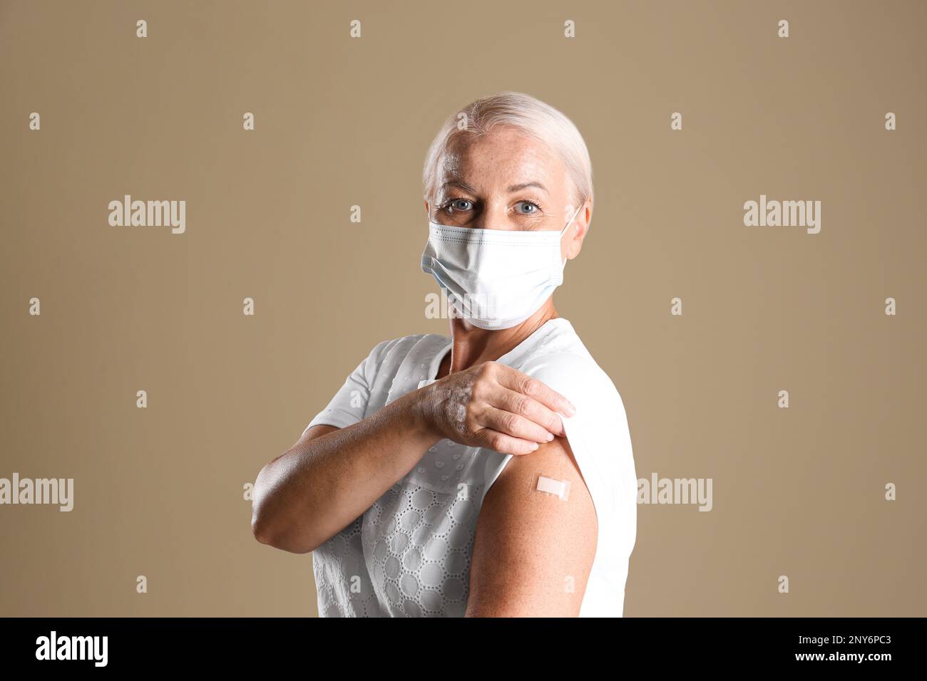 Mature woman in protective mask showing arm with bandage after vaccination on beige background Stock Photo