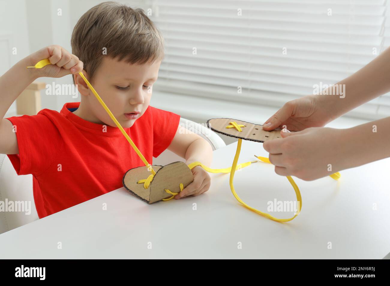 Picture of Mother teaching son to tie shoe laces using training cardboard template at white table, closeup ...