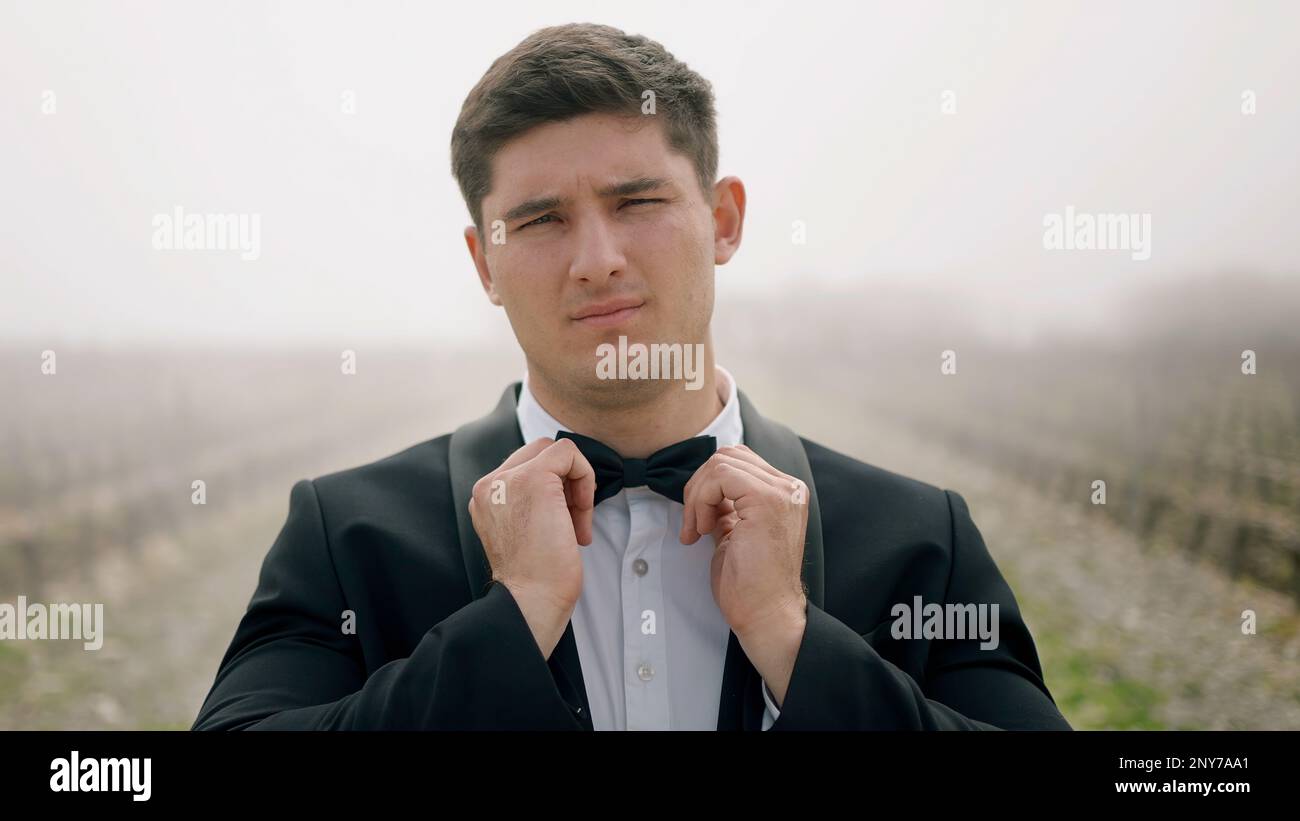 Young man in suit touching its bow tie outdoors. Action. Man wearing tuxedo standing in the field with fog Stock Photo