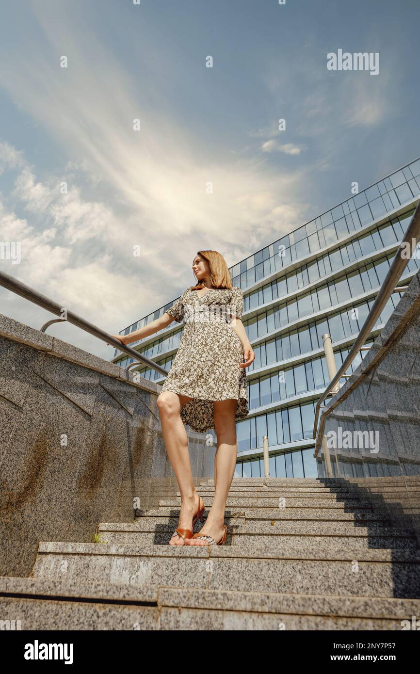 A young woman stands atop a majestic staircase, her fashionable footwear and sundress contrasting with the sky and clouds above Stock Photo