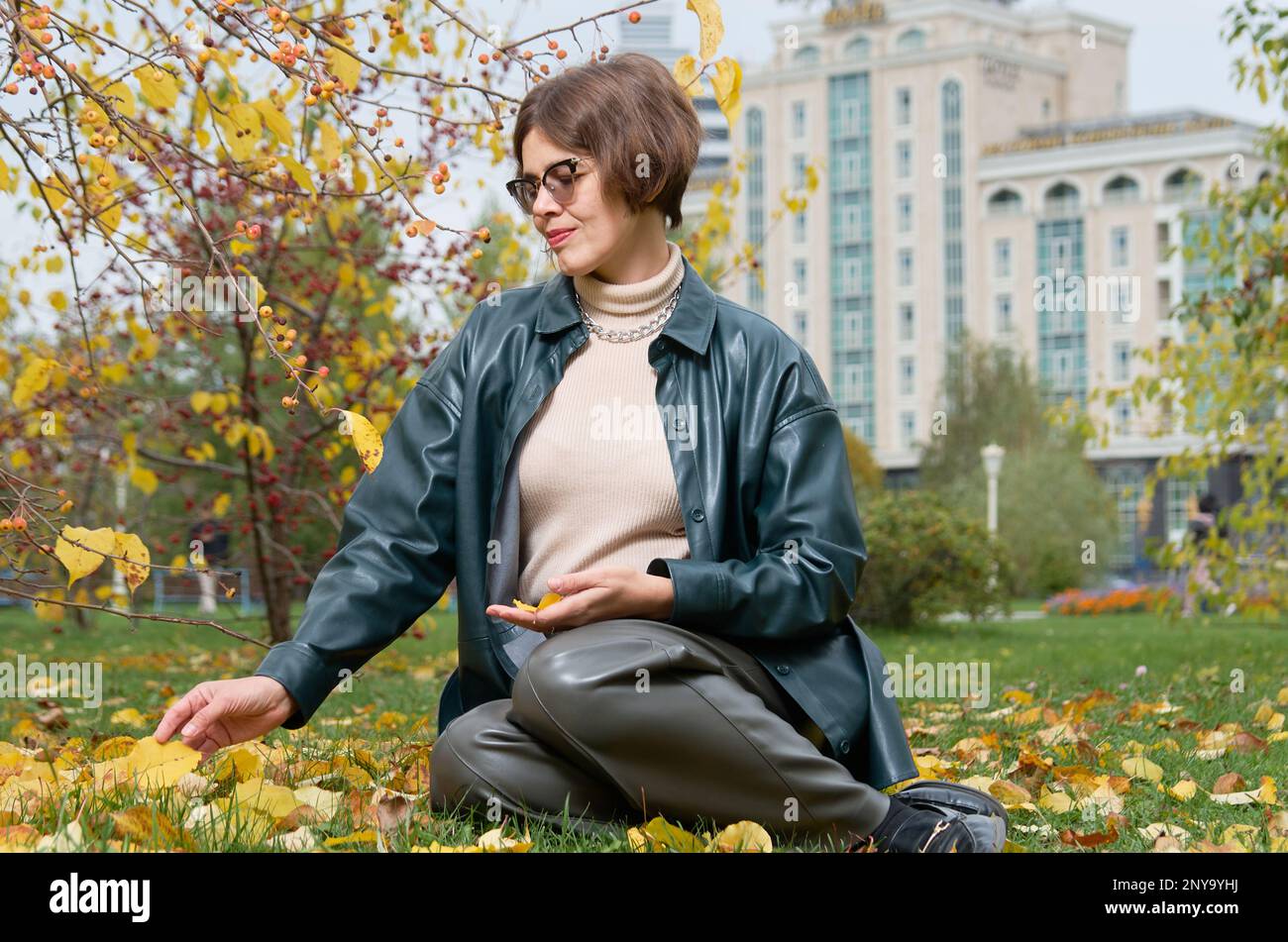 A woman in leather clothes sits on the ground among yellow leaves in an autumn park Stock Photo