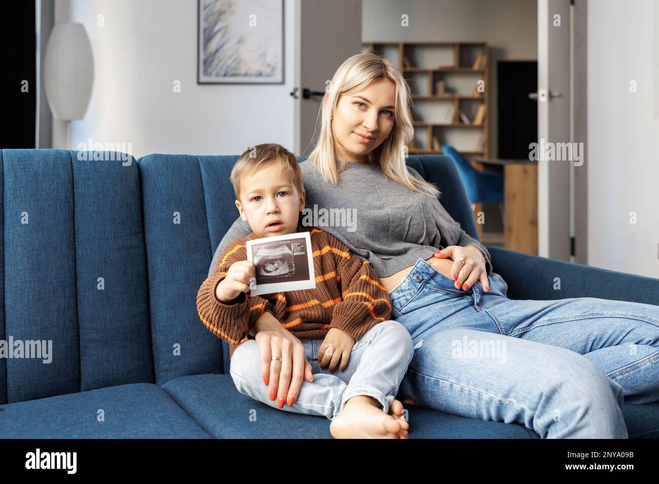 Young pregnant woman and her son sit on sofa at home and boy holds ultrasound scan in hand.They spend time together, enjoy communication feeling kicks Stock Photo