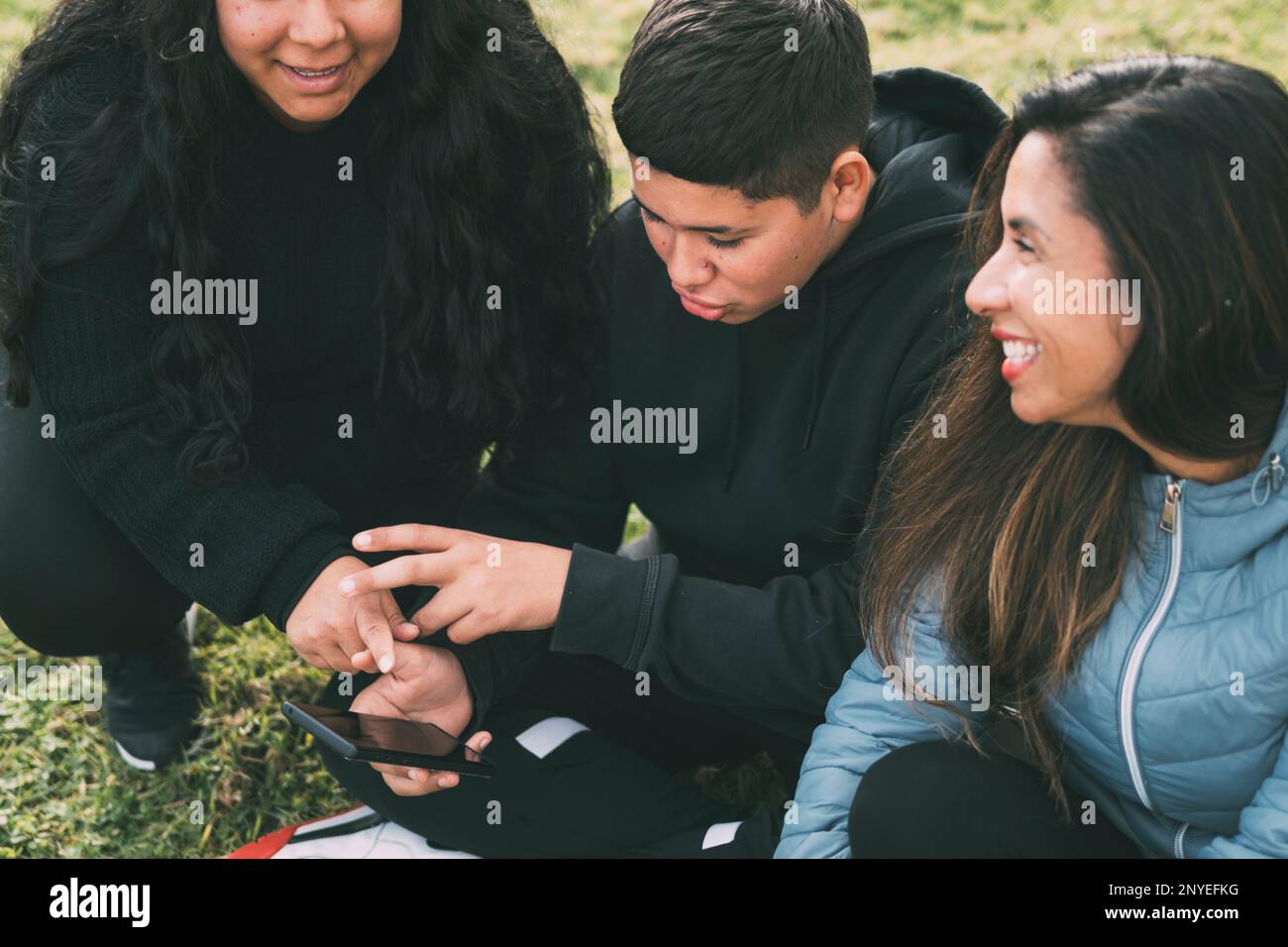 three people of hispanic-latino ethnicity sitting on the green grass of a park, with smart device. relaxed and comfortable as they entertain themselve Stock Photo
