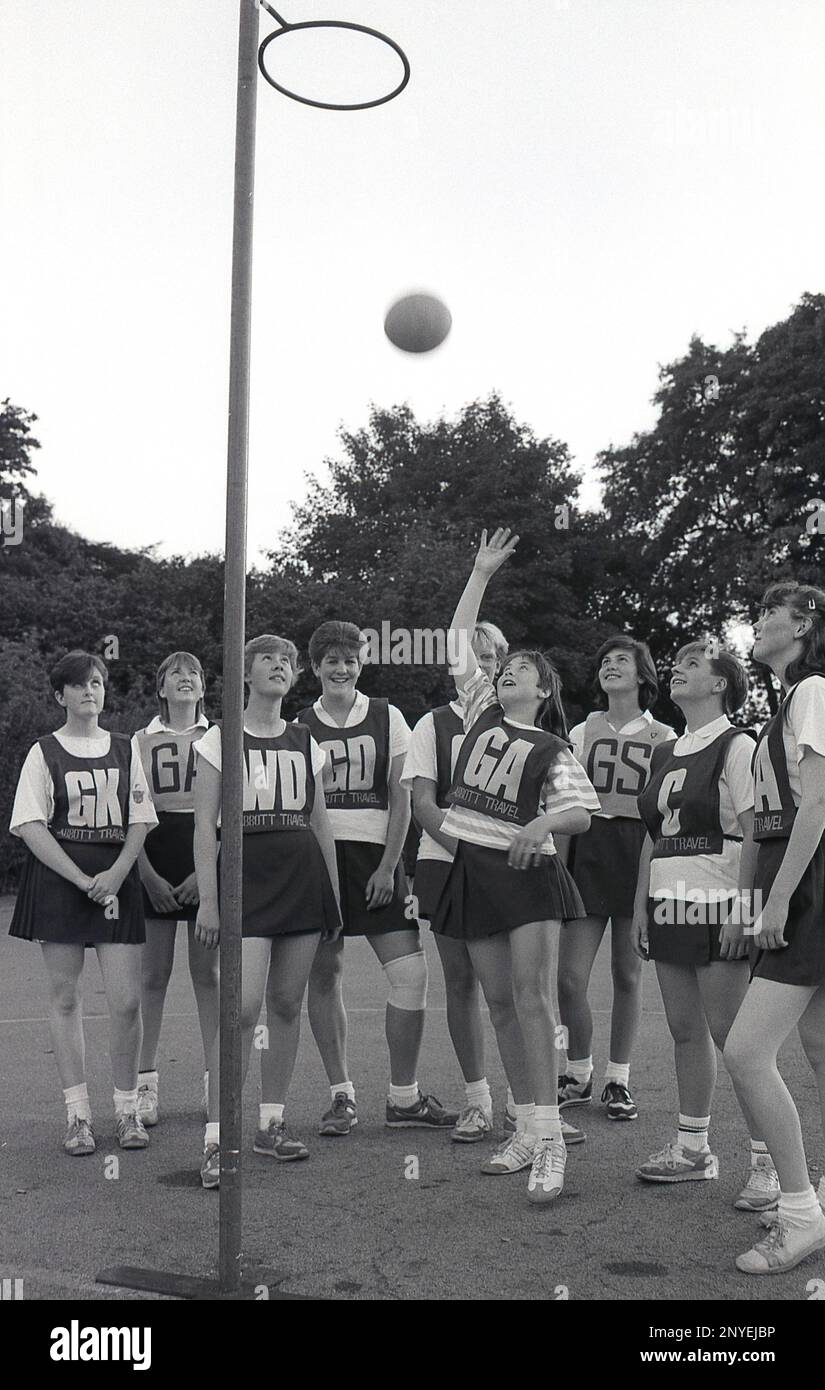 1989, netball, a schoolgirls team, with letters on their vests, standing by the post, England, UK, one of the girl's (GA) putting the ball in the raised goal ring. The vest letters stand for the seven positions in netball, i.e GA-Goal Attack. Stock Photo