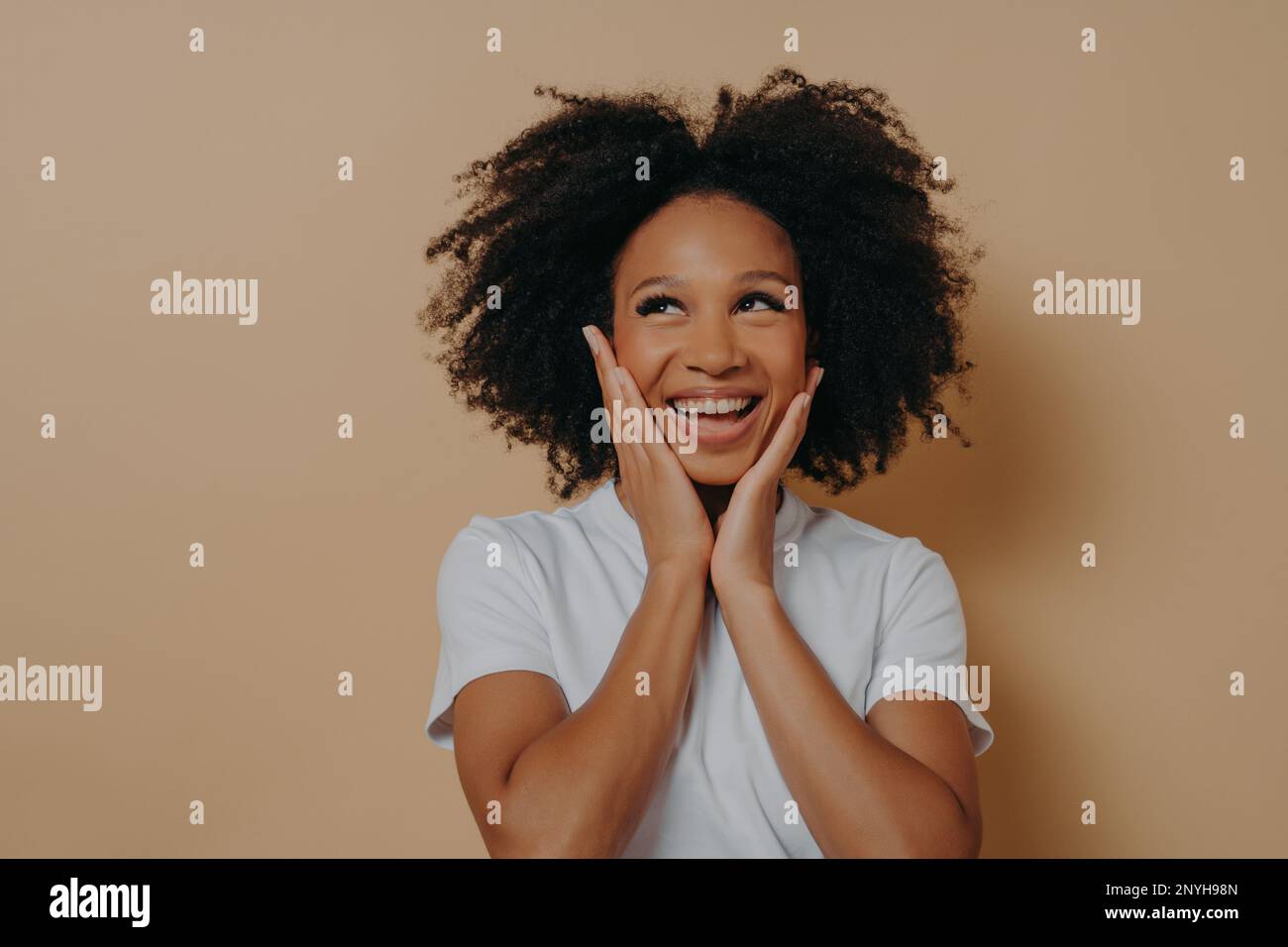 Joyful young dark skinned woman feeling happy, smiling at camera and touching cheeks with both hands, carefree african female in white tshirt expressi Stock Photo