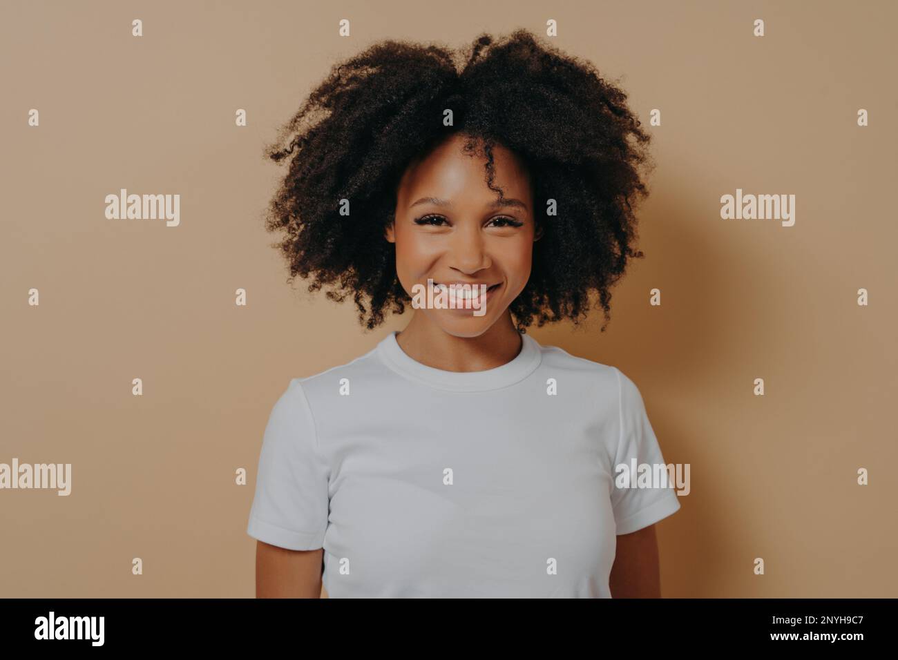 Portrait of happy young african woman with broad shining smile dressed in white tshirt posing alone against beige background, positive mixed race fema Stock Photo