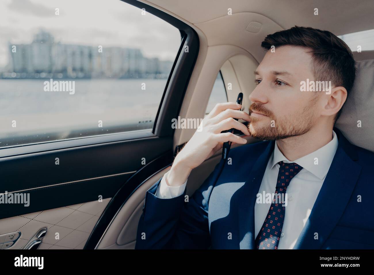 Serious male company executive in blue suit thinking of making important serious decision, weighting in risks and profits, holding his phone against h Stock Photo