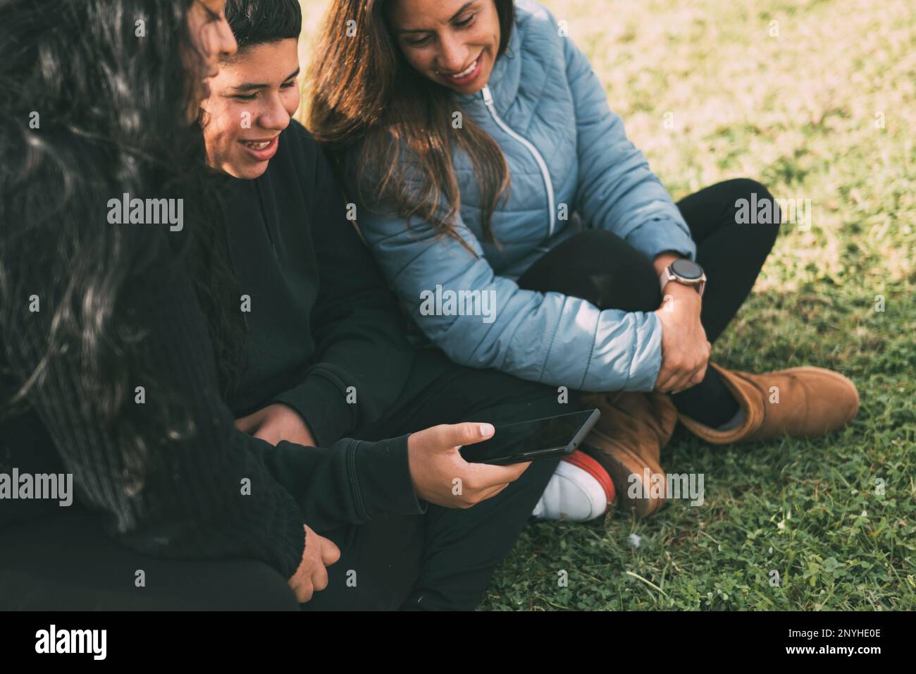Hispanic family spending quality time together in a local park on a beautiful, sunny day. A teenage boy is sitting on the grass, holding his smartphon Stock Photo