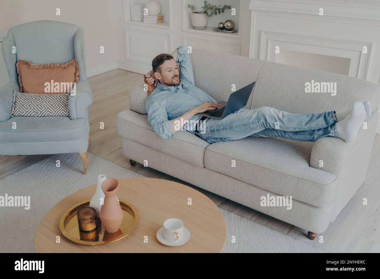 Young handsome successful businessman working remotely at home, compiling data with his notebook laptop while lying on couch with head resting on hand Stock Photo