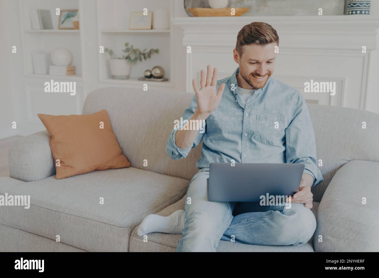 Young handsome casually dressed man waving his hand while speaking with family online using notebook, happy to have conversation with relatives, sitti Stock Photo