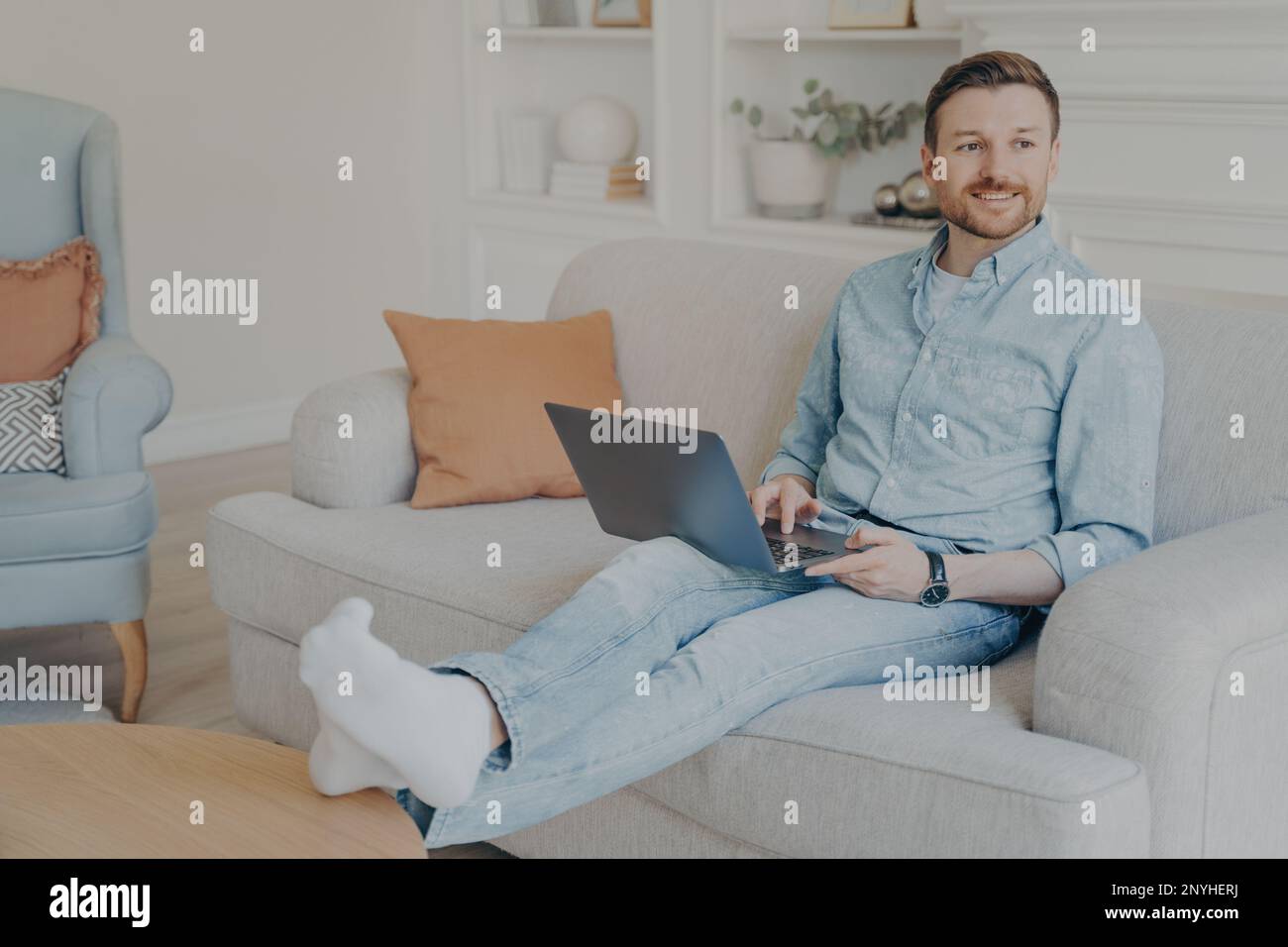 Young man with stubble surfing web on his laptop while sitting on couch, resting legs on coffee table, relaxing after hard work session on project, ta Stock Photo