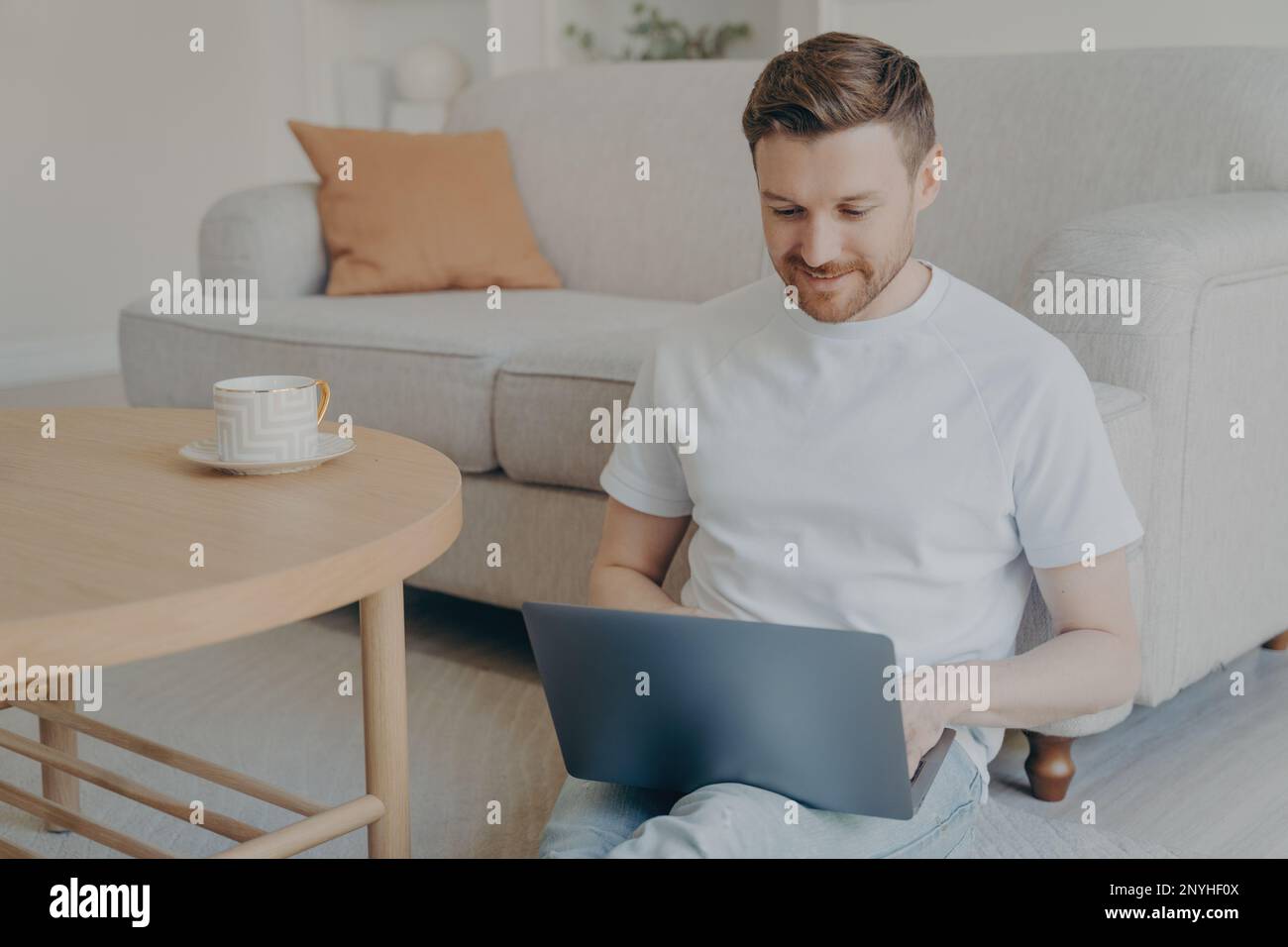 Attractive caucasian bearded freelancer man having his morning cup of coffee while sitting on carpeted floor in modern living room interior with lapto Stock Photo