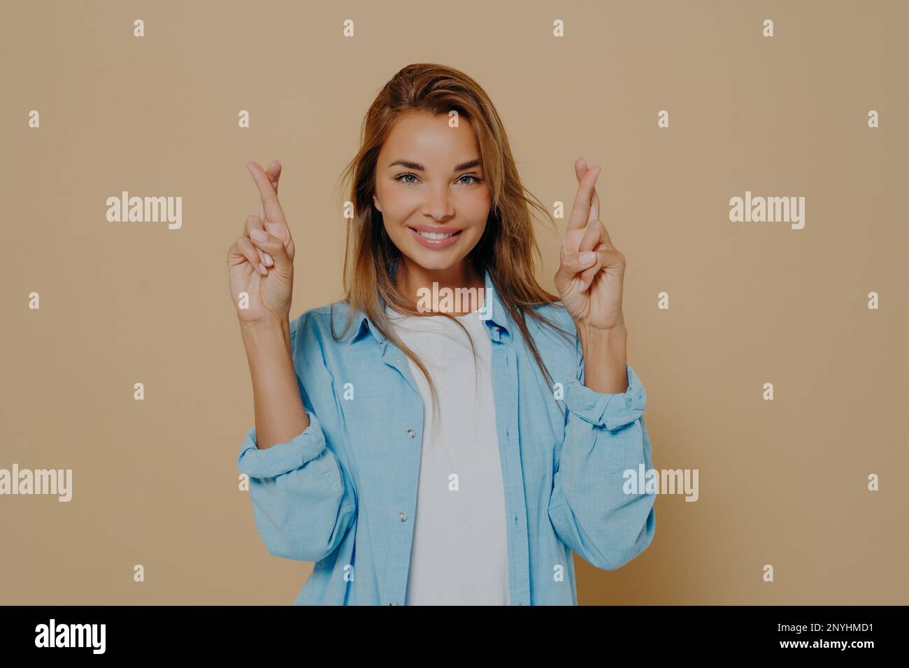 Positive superstitious young lady with beautiful smile holding her fingers crossed, making believer gesture, hoping for good luck and fulfillment of d Stock Photo