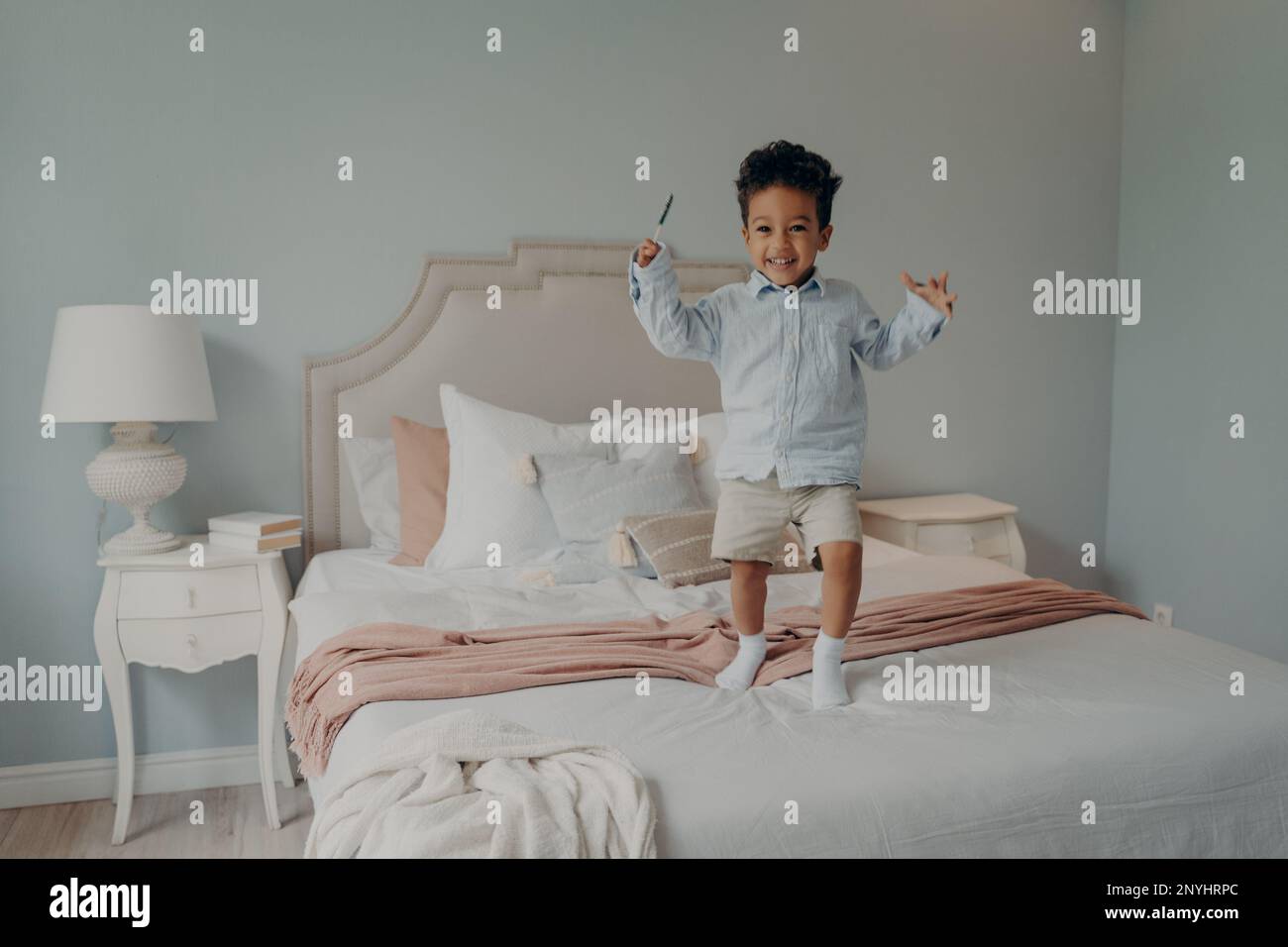 Joyful small mixed race boy happy kid dressed in blue shirt and beige shorts jumping on big comfy bed with candy, expressing his happiness with shinin Stock Photo