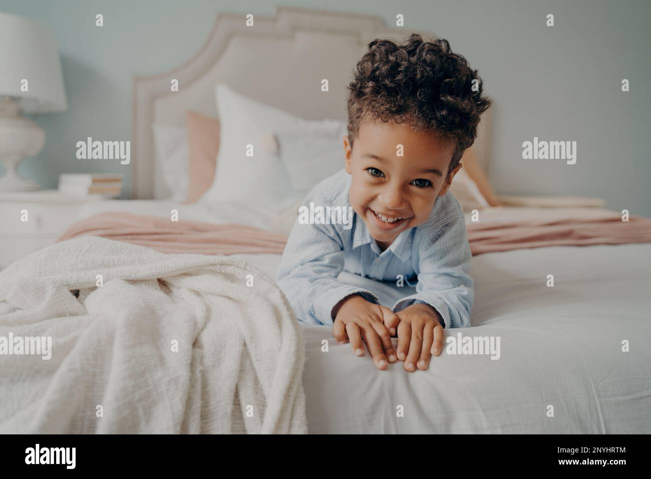 Portrait of happy curly mulatto boy smiling at camera while laying on cozy bed, relaxing after fun playing session at home, adorable preschool child e Stock Photo