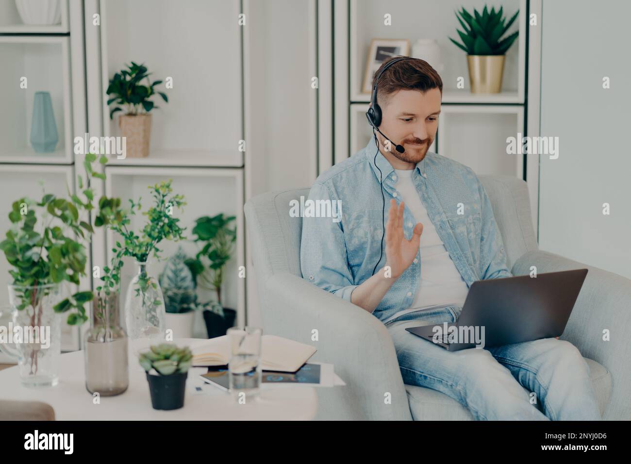 Happy guy in headset waving hand at laptop webcam while sitting in ...