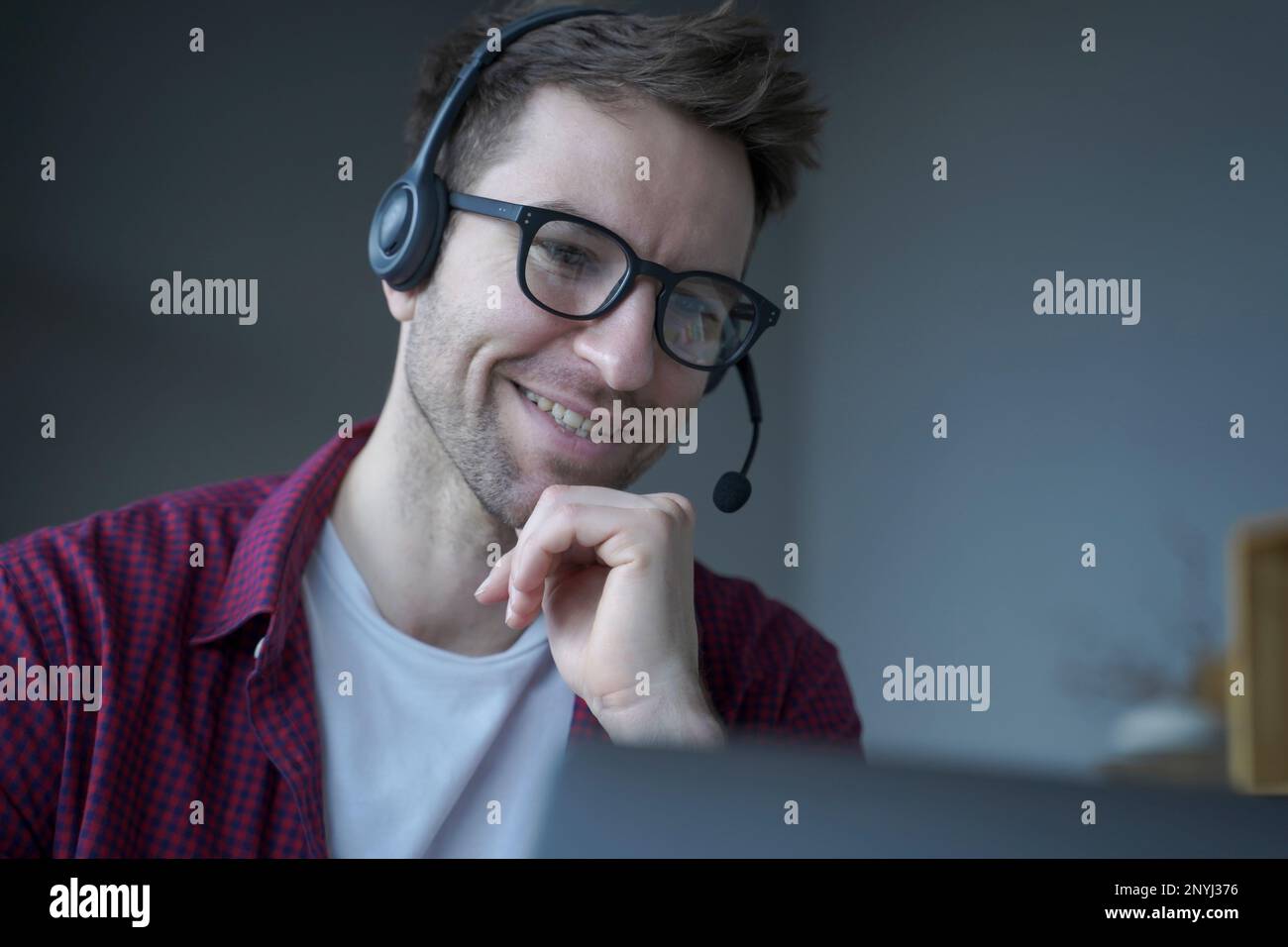 Cheerful man German speaking callcenter consultant in headset with mic works remotely from home, politely smiling while communicates online via laptop Stock Photo