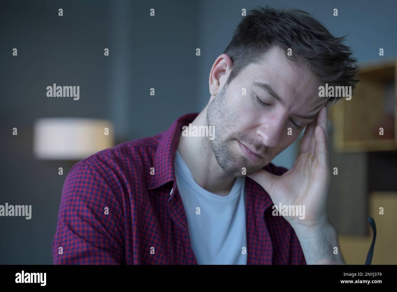 Tired European freelancer man sitting at home office working remotely online, holding one side of his head while eyes closed feeling stressed, sufferi Stock Photo