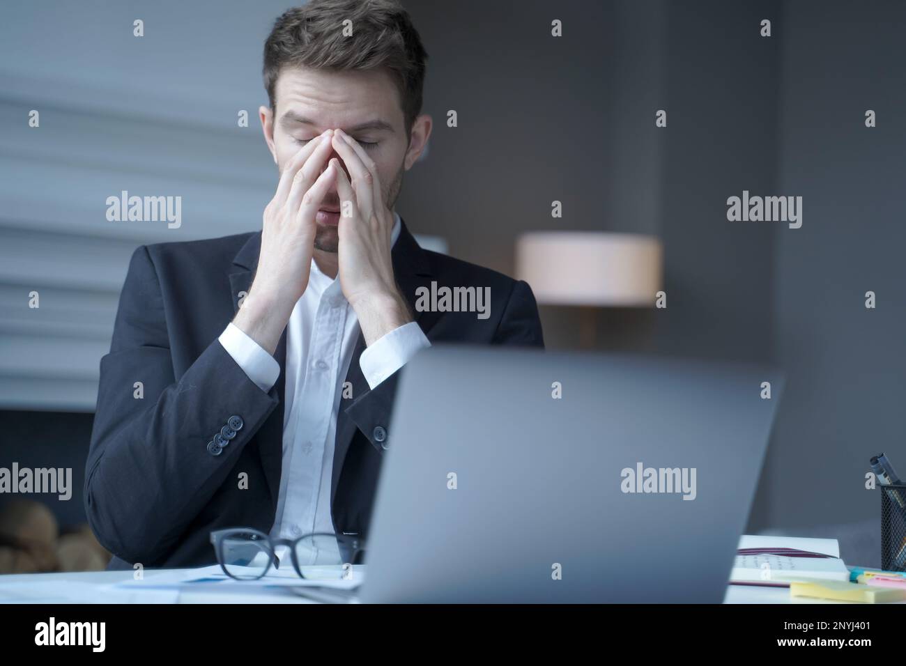 Overworked male entrepreneur sits at home office while took off glasses massages bridge of his nose with eyes closed, exhausted man suffering from hea Stock Photo
