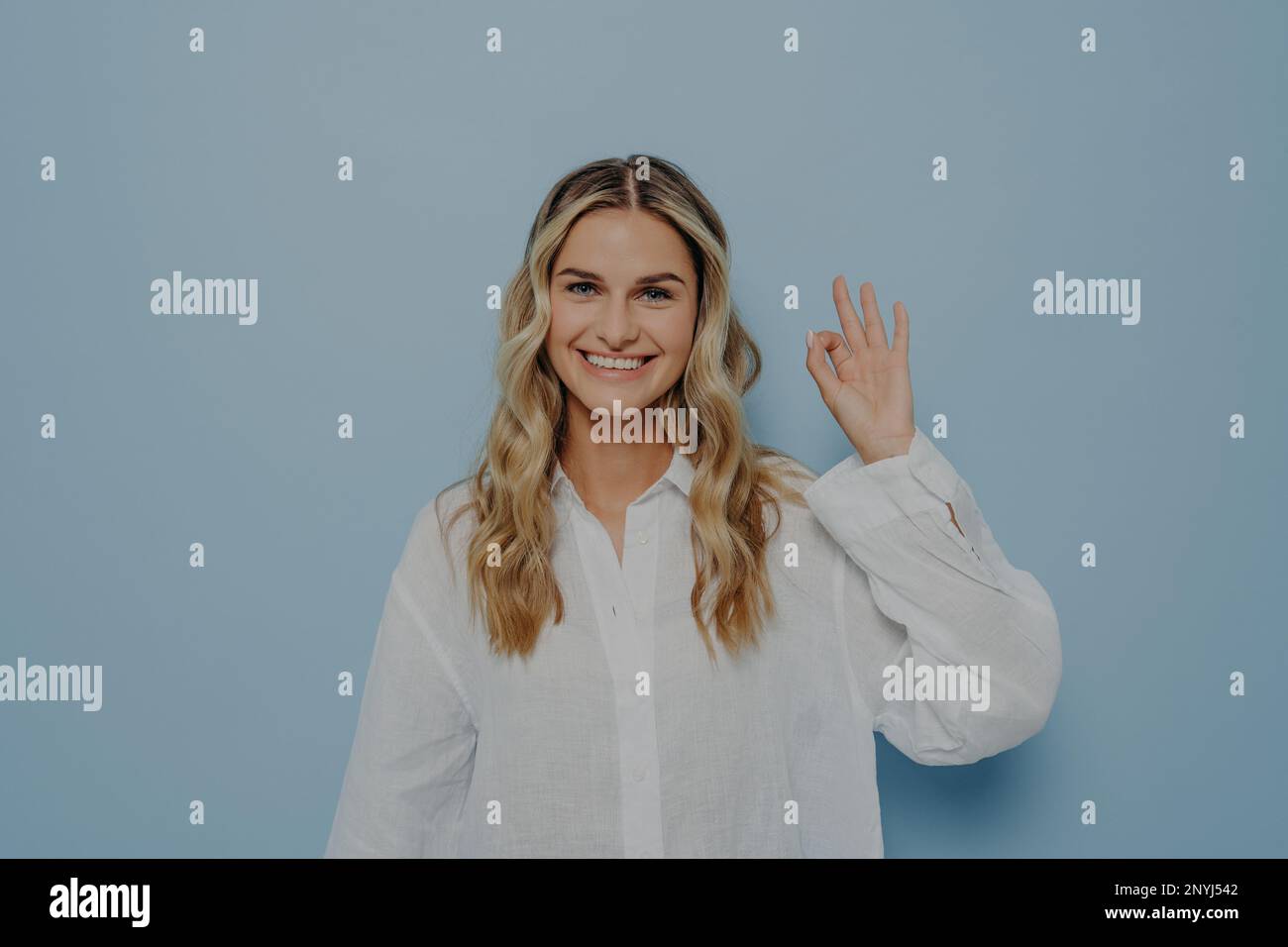Joyful young blonde woman in white shirt making ok gesture with her hand in air, showing that everything is alright, signalling there is no problem wh Stock Photo