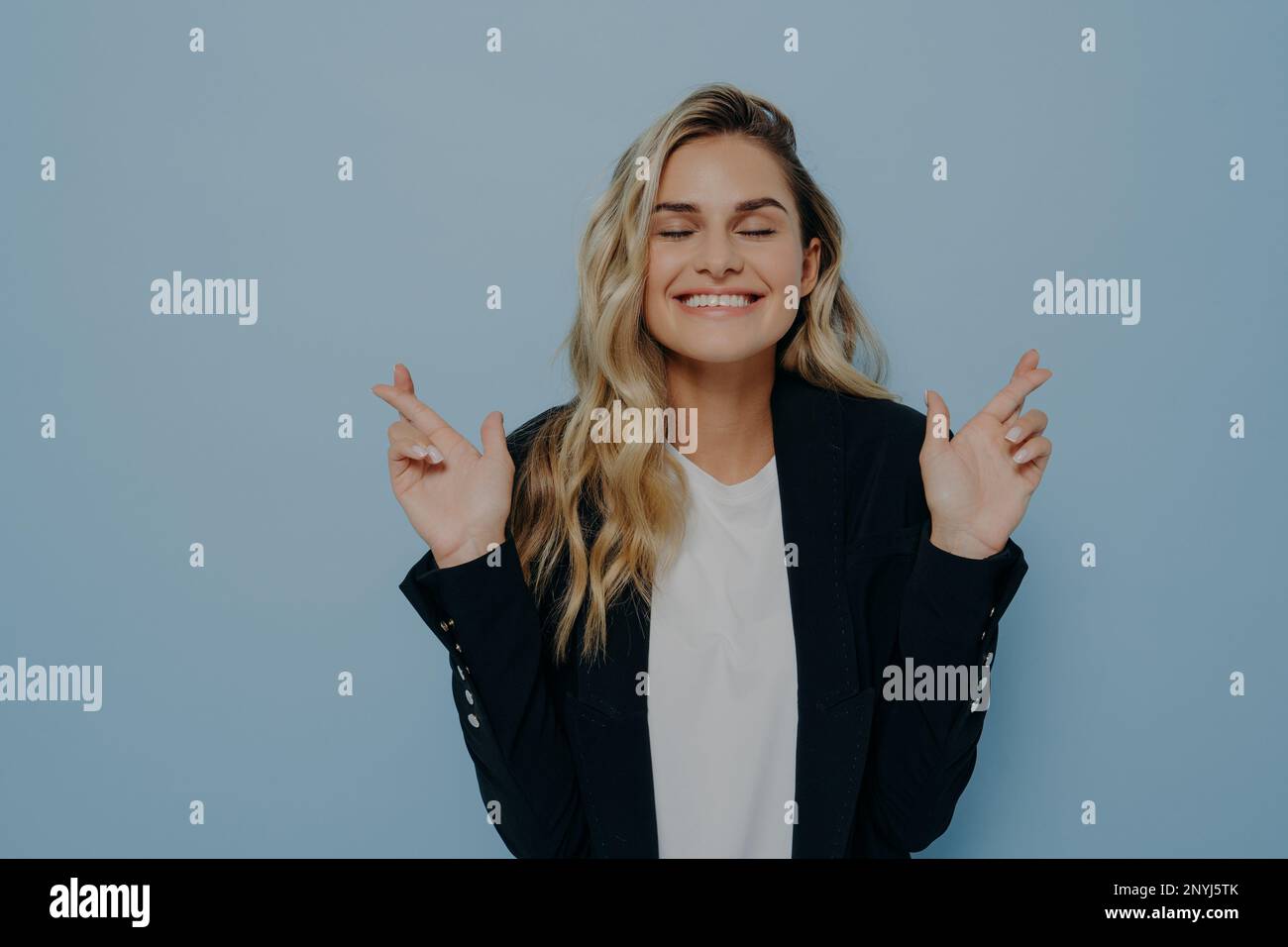 Superstitious young student girl with blond hair crossing fingers with both hands, invoking good luck before important exam or event, hopes for victor Stock Photo