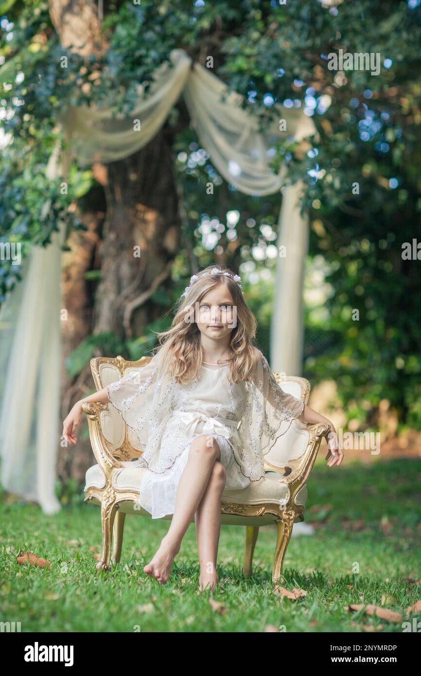 Little girl barefoot in white dress and flower crown sitting on antique ...