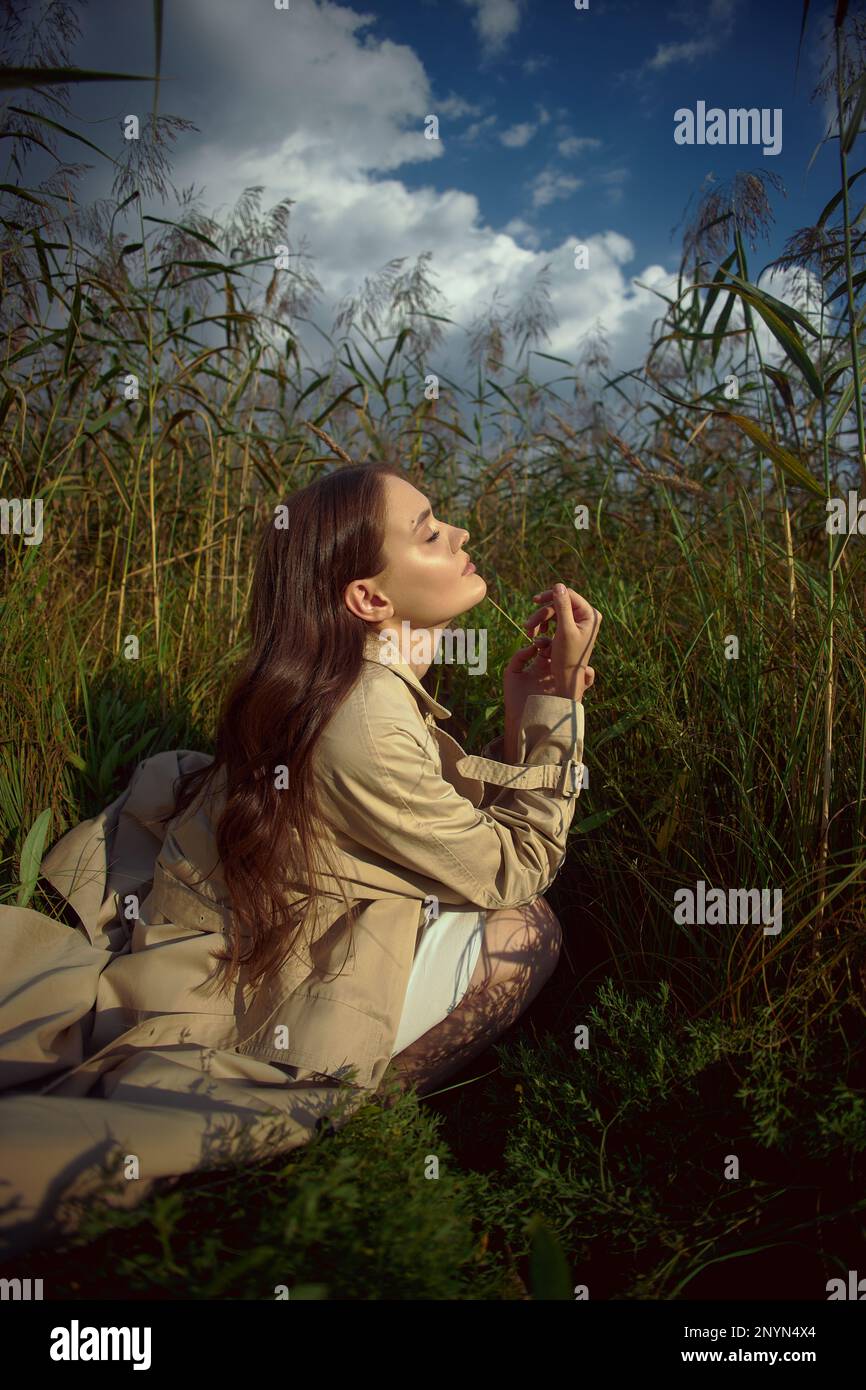 Dreamy gentle woman in a beige raincoat in nature. Romantic young woman in thick tall grass in field. Natural beauty, enjoyment of life Stock Photo