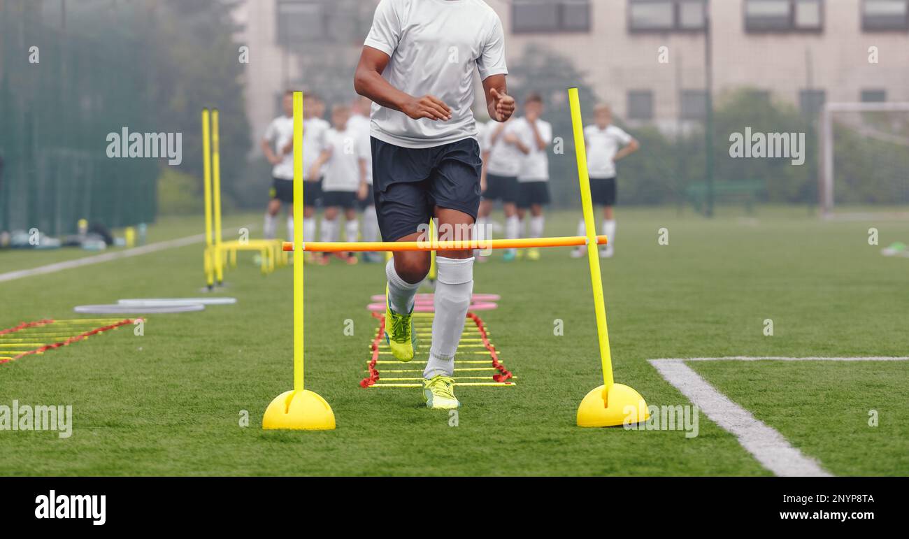 Teenage african player running at practice drill. Soccer teenager jumping over training ladder and hurdles. Football teeam in blurred background Stock Photo