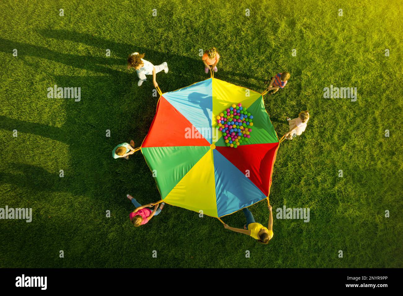 Group of children and teachers playing with rainbow playground parachute on green grass, top