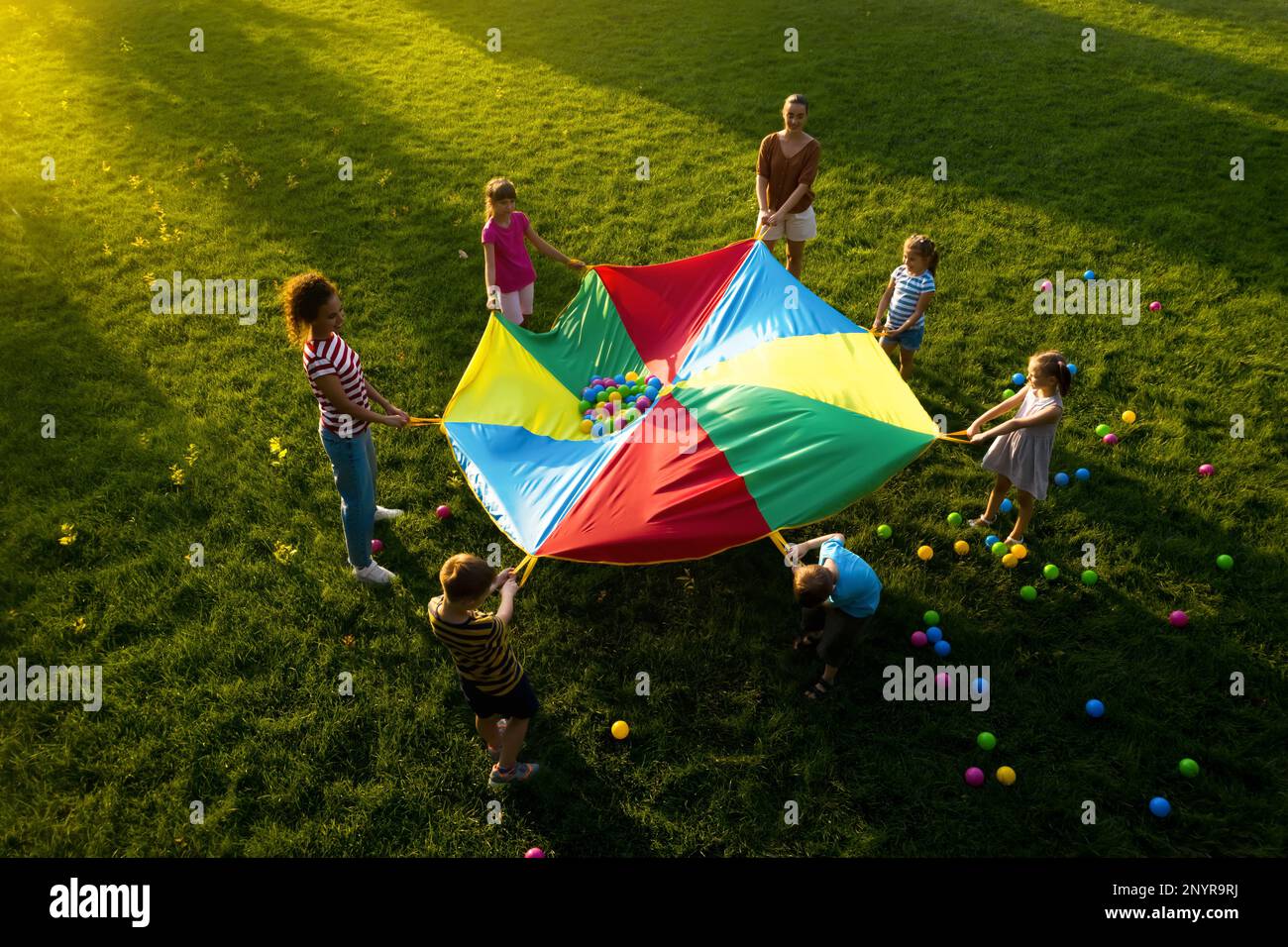 Group of children and teachers playing with rainbow playground parachute on green grass, above