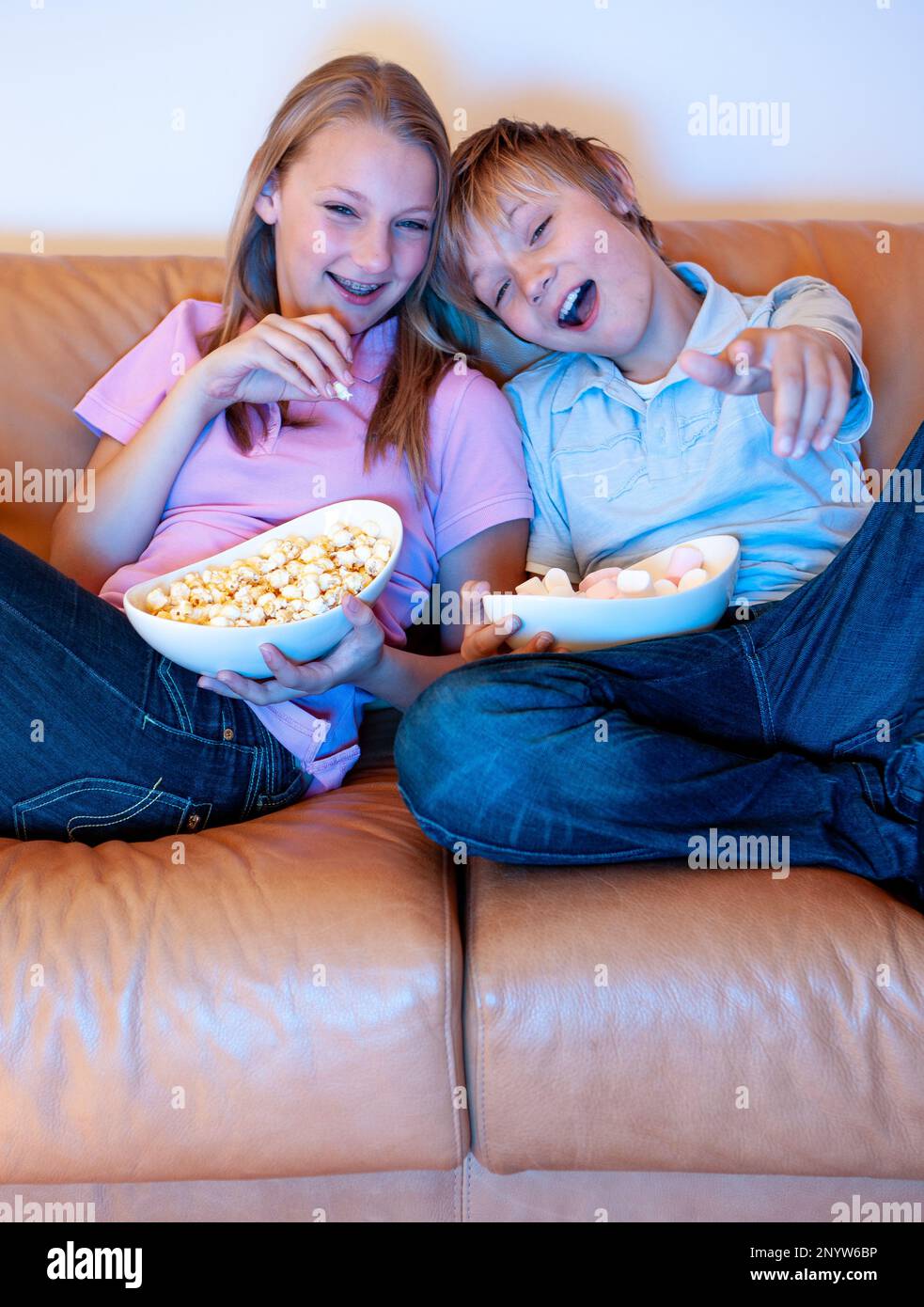 Family Life, Watching TV. Candid and carefree siblings enjoying sharing snacks and their favourite television show. From a series of related images. Stock Photo