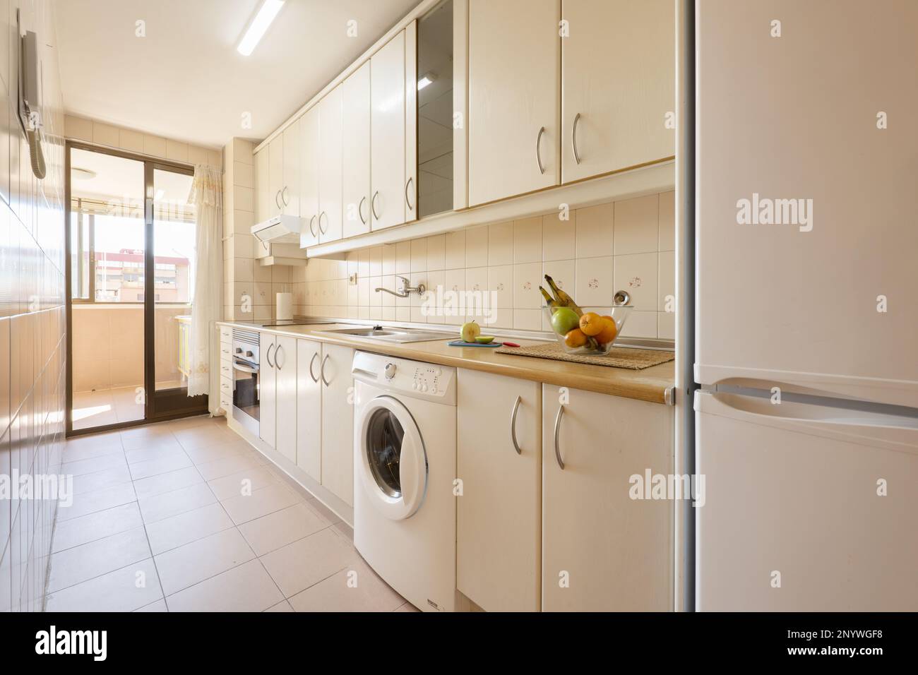 Furnished kitchen in a stereo room on one side of a wall with light wood furniture, integrated white appliances and exit to a terrace with a glass doo Stock Photo