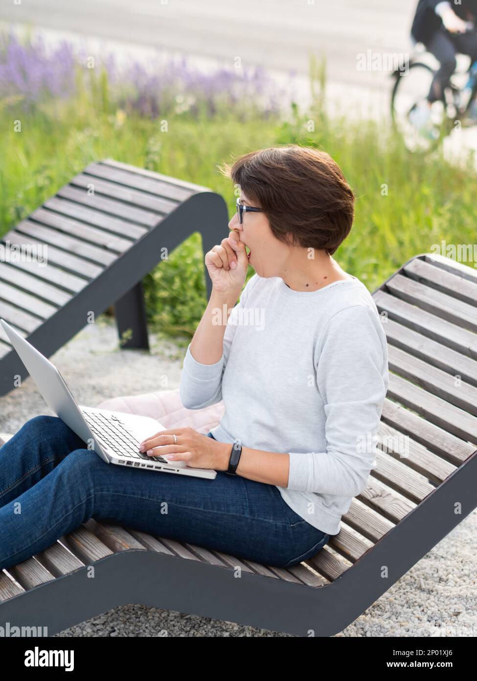 Woman sits with laptop on urban park bench. Freelancer yawns at work. Student learns remotely from outdoors. Modern lifestyle. Stock Photo