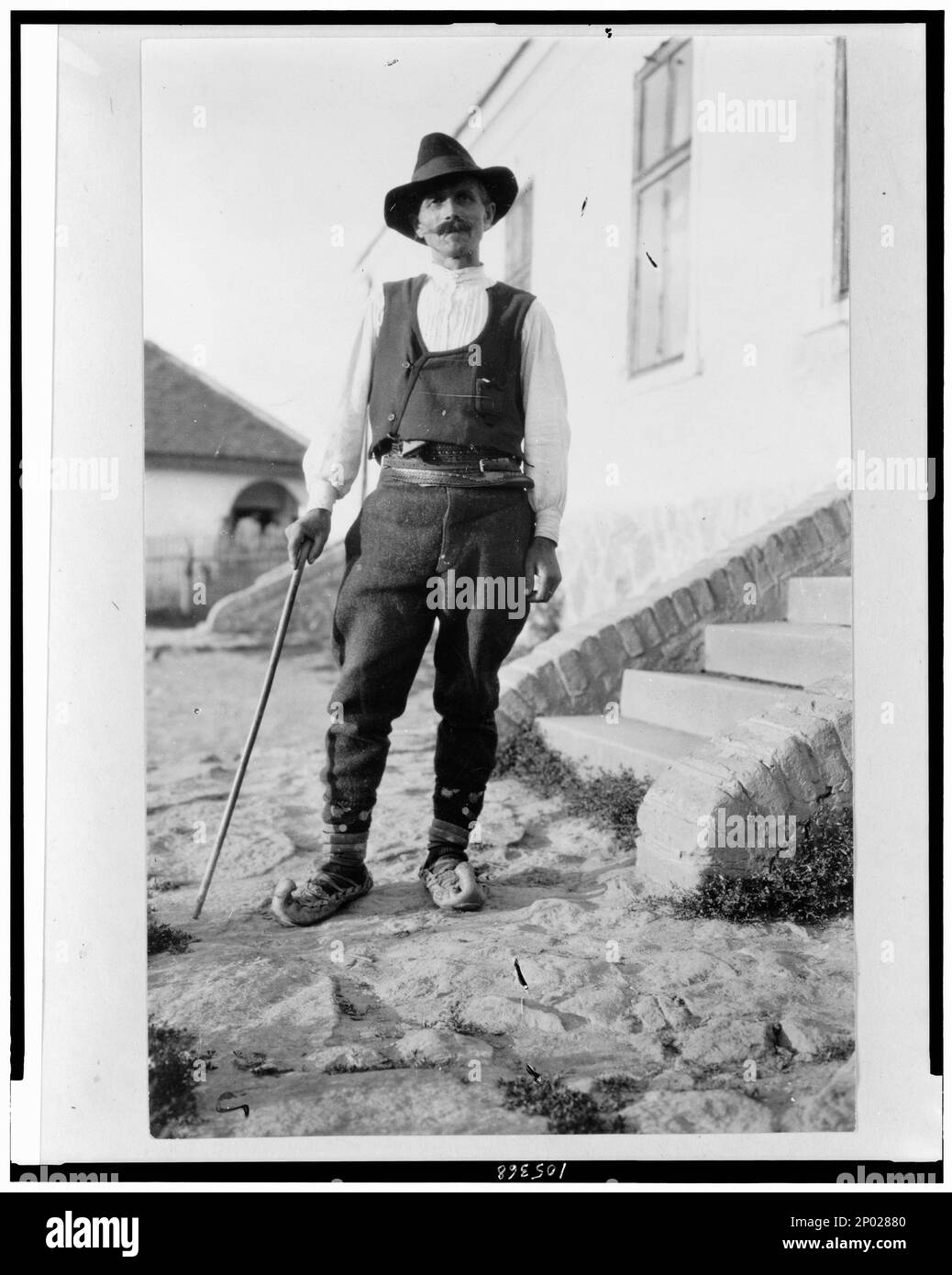 Serbian man wearing hat, vest, belted pants, and pointed toed shoes with cane standing outside of building in Yugoslavia. Frank and Frances Carpenter Collection, No. 963, Serbs,Clothing & dress,Yugoslavia,1880-1930. Stock Photo