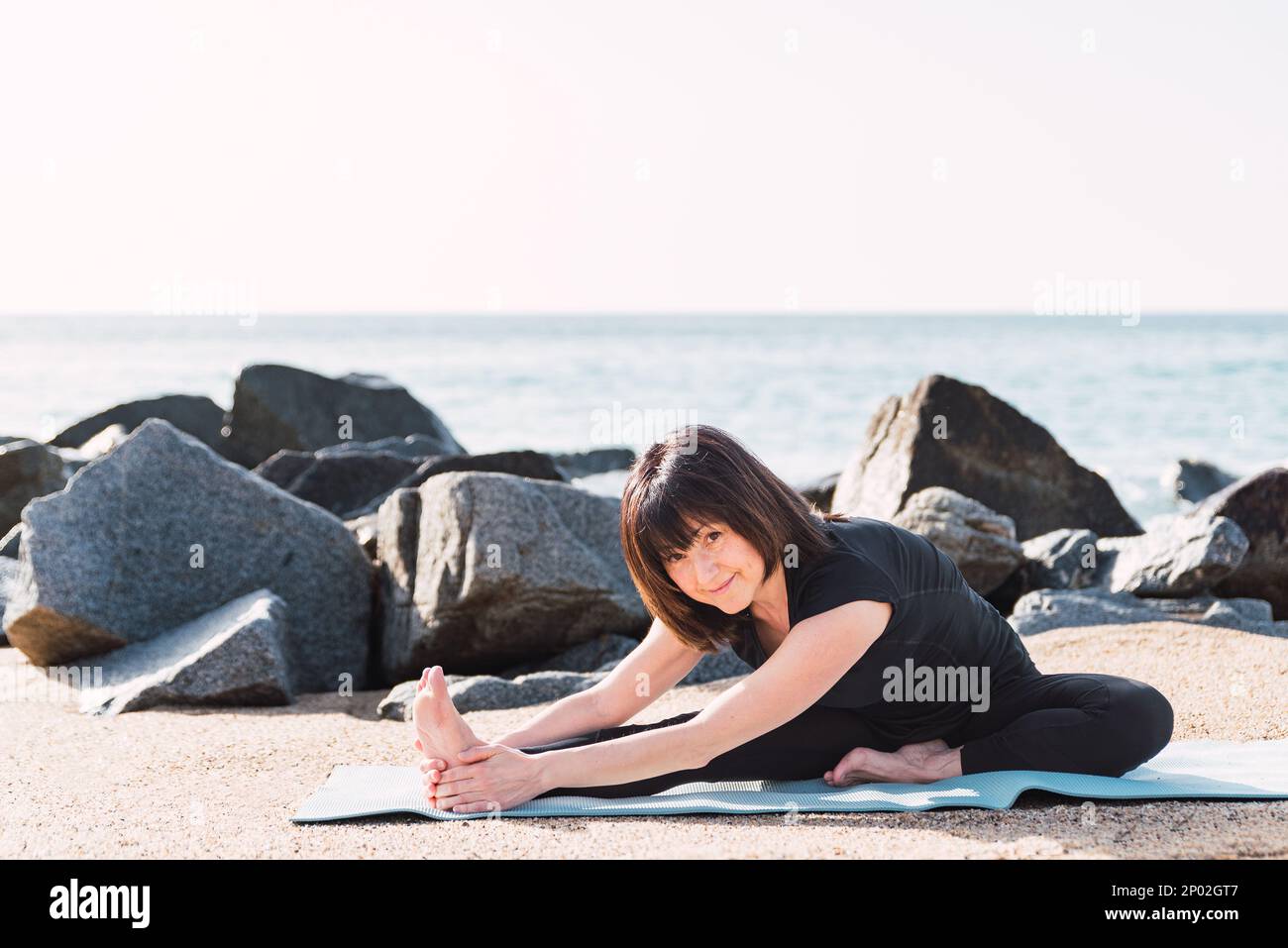 Cheerful barefoot female in sportswear sitting on mat in Head to Knee posture. She is stretching legs while doing yoga on shore near sea in sunny day and looking at camera Stock Photo
