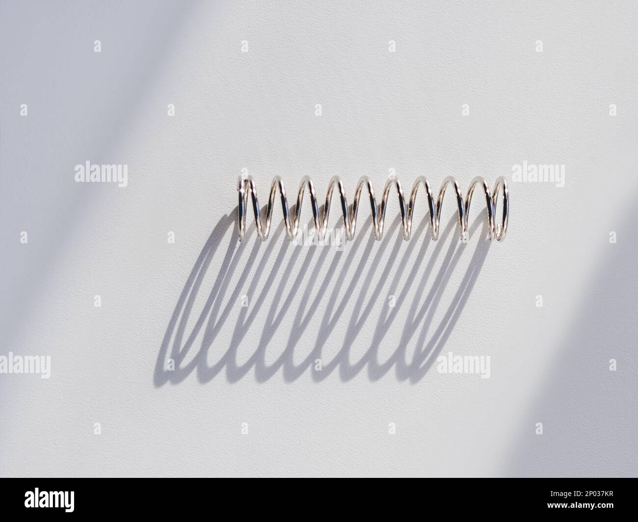 Metal mainspring on white background. Flexible chrome tube. Corrugated object on geometrical banner with copy space on hard sunlight and shadow. Stock Photo