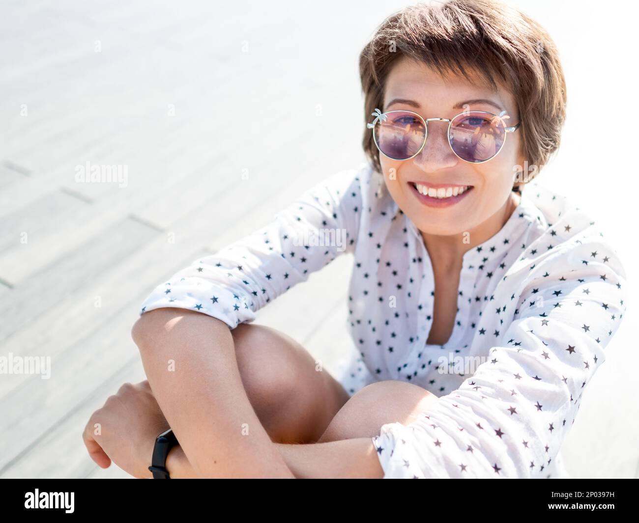 Woman with short hair and freckles poses in colorful sunglasses. Smiling woman at open wooden scene of urban park. Summer vibes. Sincere emotions. Stock Photo