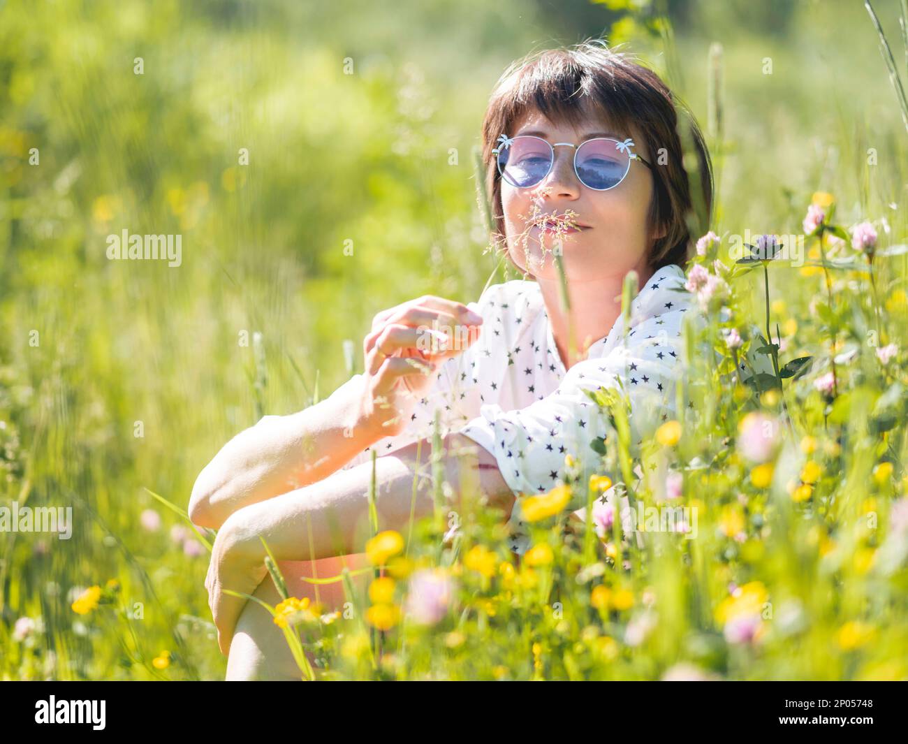 Woman in colorful sunglasses, enjoys sunlight and flower fragrance on grass field. Summer vibes. Relax outdoors. Self-soothing. Stock Photo